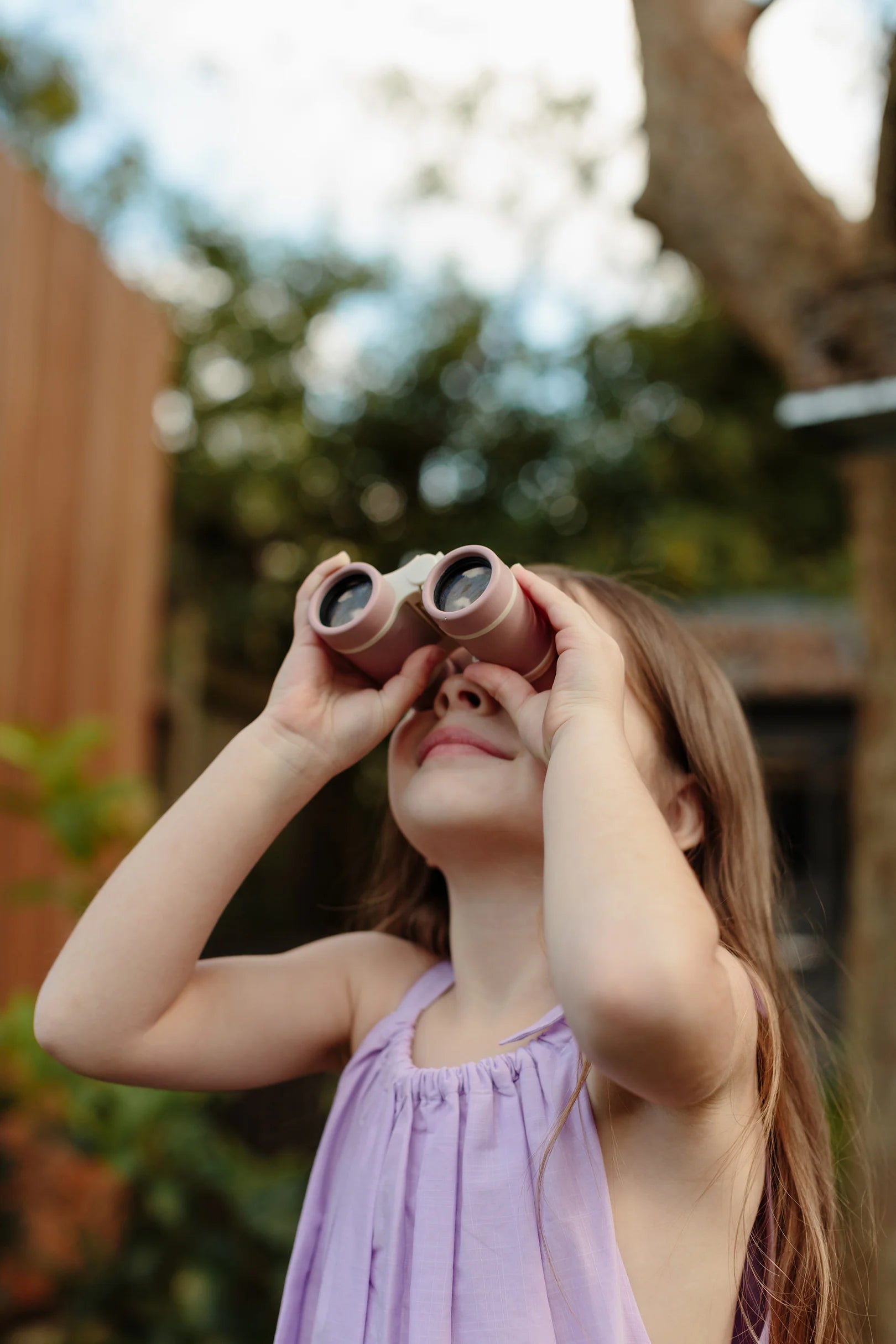 A young girl with long hair, wearing a light purple sleeveless dress, is looking up through a pair of LITTLE DROP's Kids Explore Binoculars Rose. She stands outdoors, participating in a scavenger hunt, with greenery and a wooden fence blurred in the background.