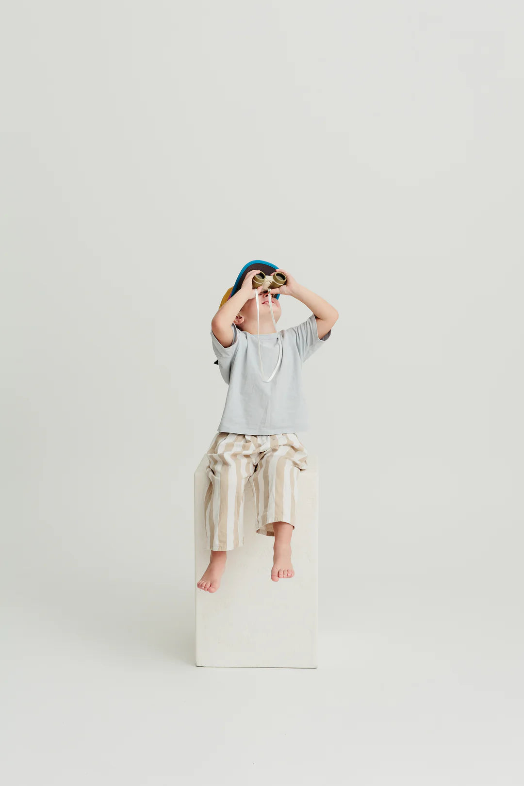 A child sits on a white block against a plain background, wearing a grey shirt, striped beige pants, and a colorful hat. With LITTLE DROP's Kids Explore Binoculars Moss held to their eyes, they look upward as if on a scavenger hunt.