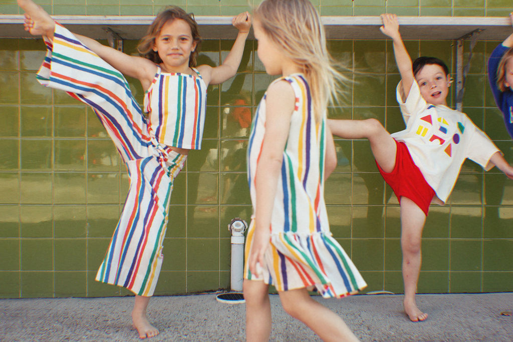 Three children in colorful, striped outfits embrace summer fun energetically by a green tiled wall. Two kids kick up their legs playfully, while one proudly shows off her GOLDEN CHILD Kaleidoscope Tank Top.