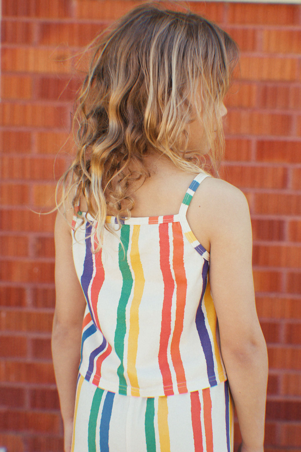 A young girl with wavy blonde hair faces a brick wall, embodying summer fun in her sleeveless Kaleidoscope Tank Top and dress by GOLDEN CHILD, featuring colorful vertical stripes in red, yellow, green, and blue.