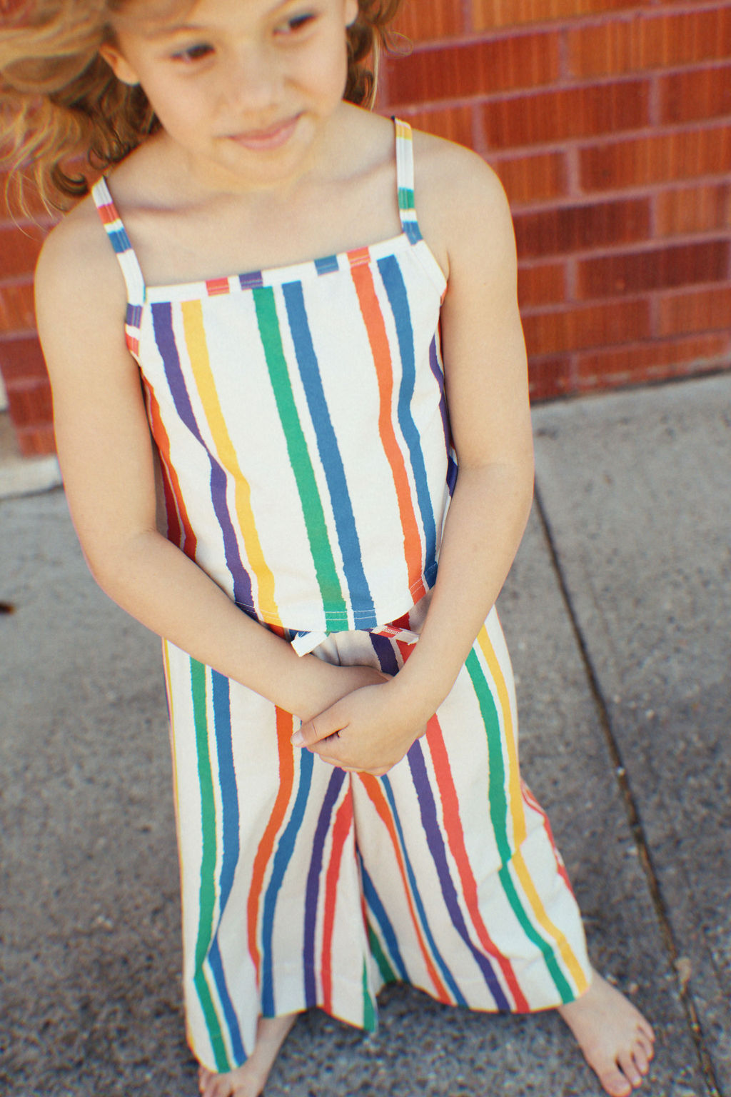 A barefoot child with slightly curly hair embodies summer fun, wearing GOLDEN CHILD's The Kaleidoscope Tank Top, which boasts multicolored vertical stripes. The scene is set against a red brick wall and concrete pavement.