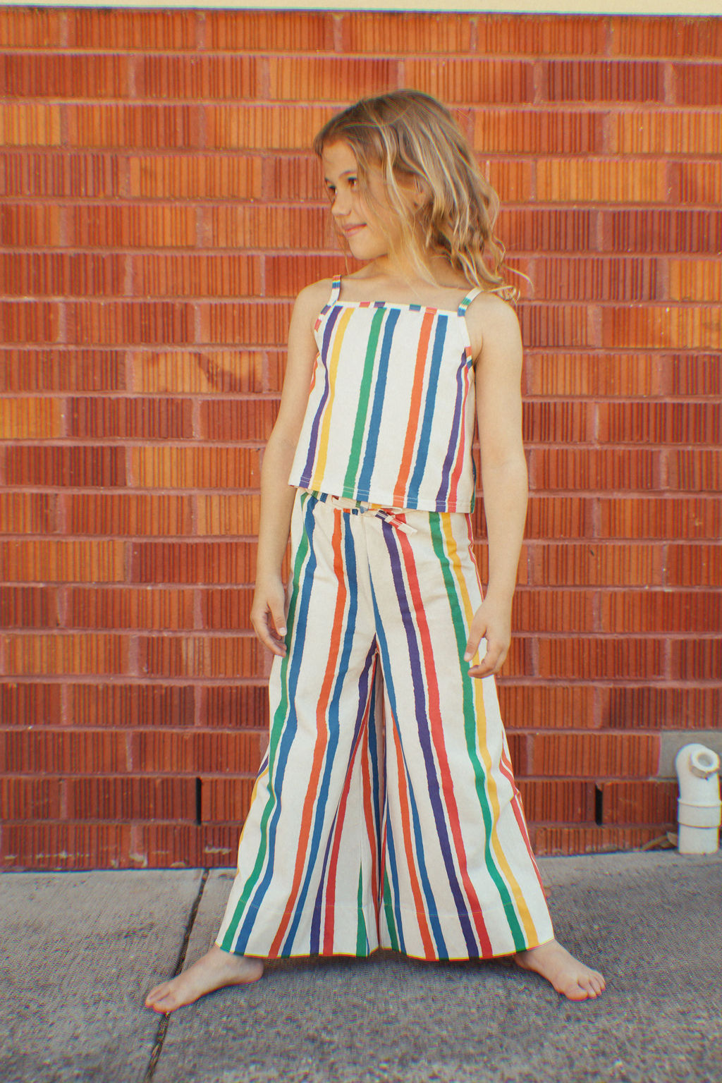 A young girl stands barefoot against a brick wall, wearing GOLDEN CHILD's The Kaleidoscope Tank Top and matching wide-leg pants. With her hands on her hips, she exudes summer fun, looking content and confident.