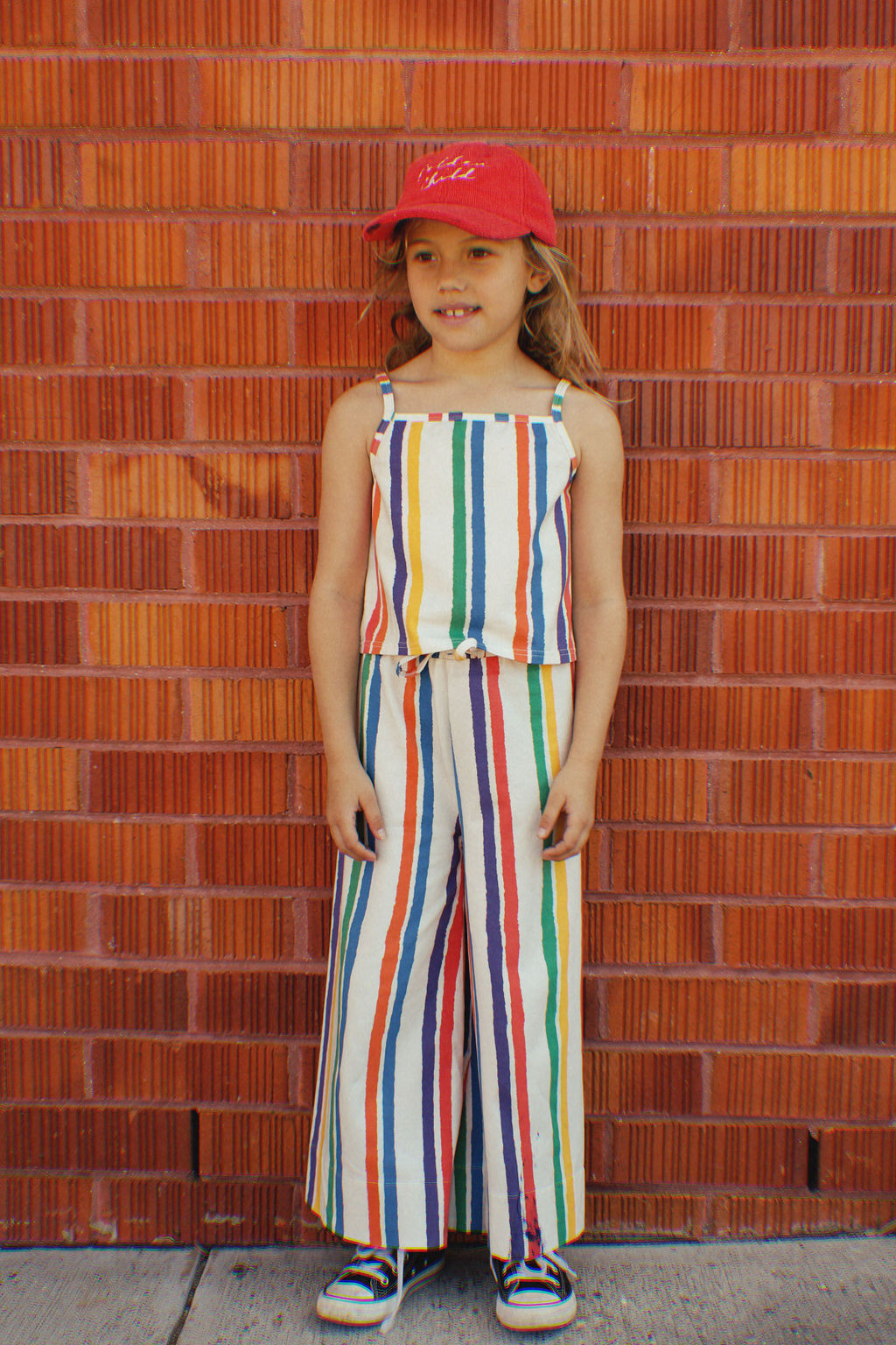A young girl stands by a red brick wall wearing GOLDEN CHILD's Kaleidoscope Tank Top and a red cap, epitomizing summer fun. Paired with black sneakers, she looks joyful and relaxed, embodying the carefree spirit of childhood adventures.