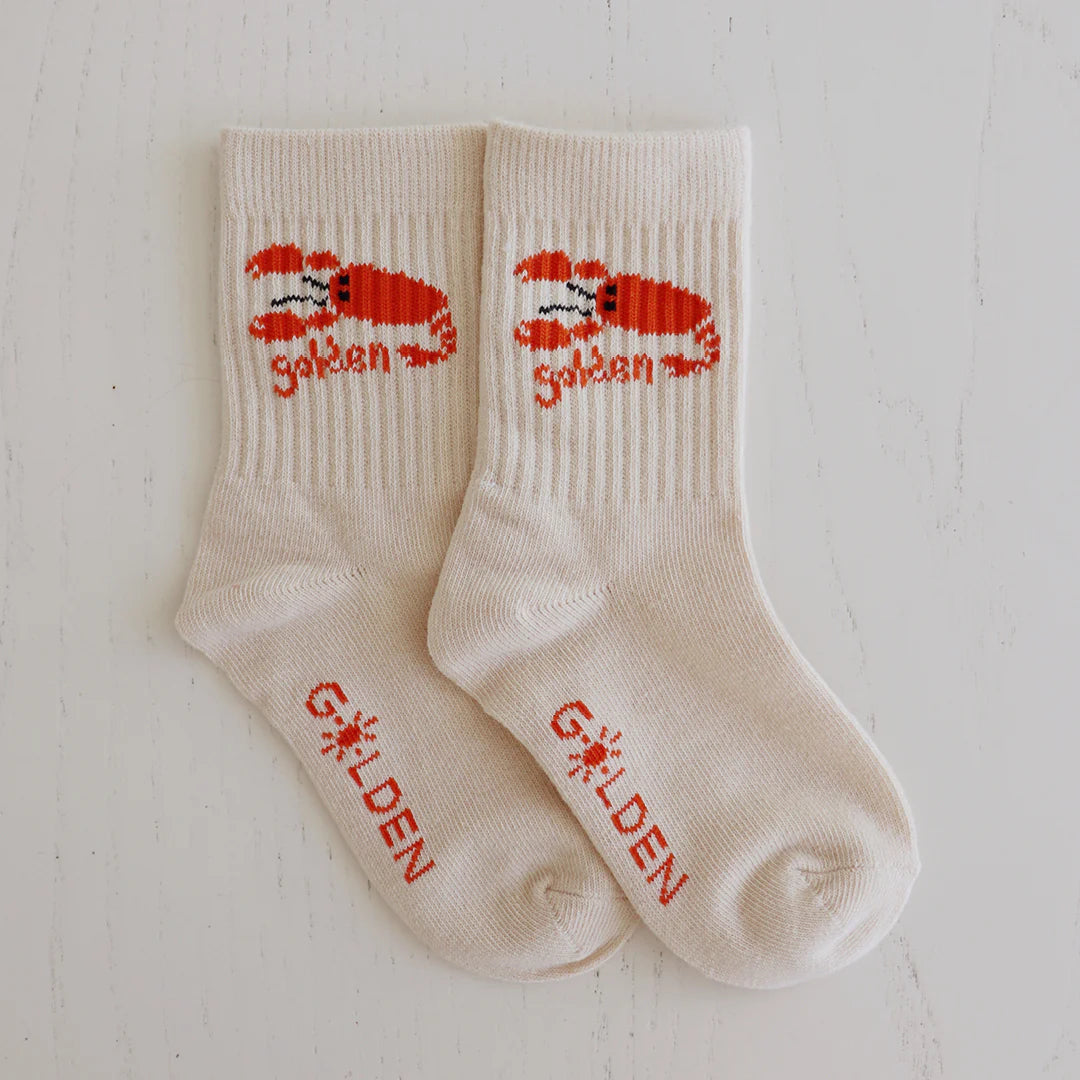 Tan ribbed socks for children from GOLDEN CHILDREN, called Lobster Socks, feature a red lobster and the GOLDEN logo on the cuff, with "GOLDEN" printed in red on the toes. The socks are displayed flat on a white background.
