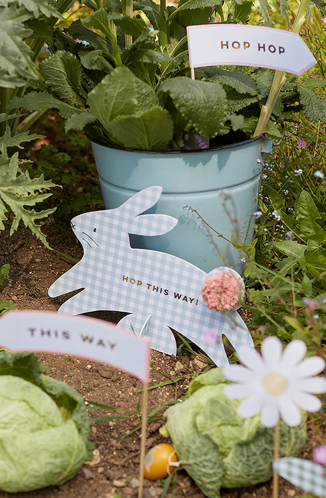 Create a charming garden scene with the Gingham Bunnies Egg Hunt Kit by MERI MERI: a blue pot of leafy greens and a whimsical paper rabbit sign "Hop This Way!" Two cabbages feature "This Way" and "Hop Hop" signs with gold foil, while a white daisy complements your perfect egg hunt setup.