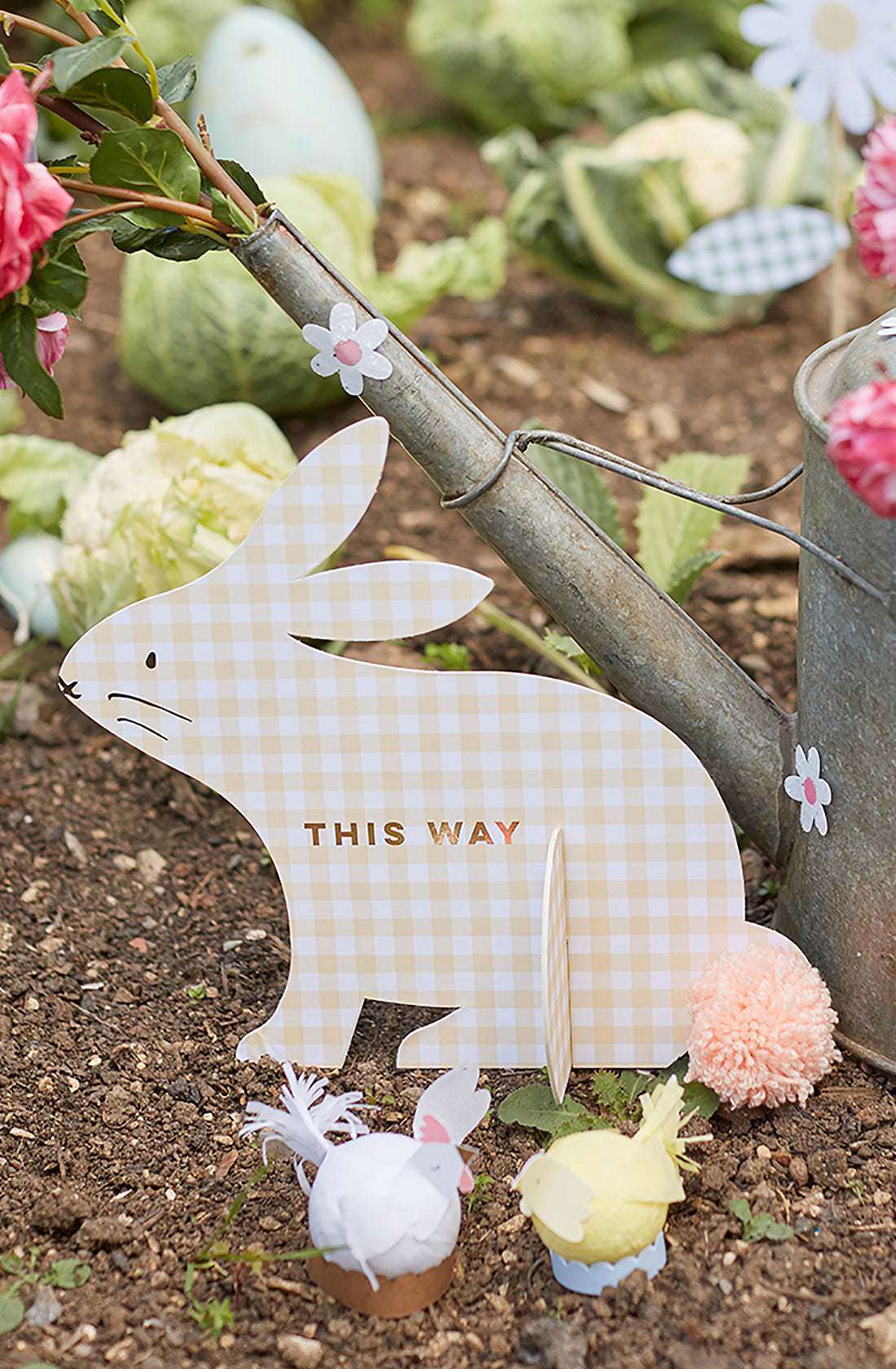 A garden scene includes a yellow checkered rabbit cutout from the MERI MERI Gingham Bunnies Egg Hunt Kit labeled with "This way" text, next to a metal watering can with gold foil detail. It’s surrounded by decorative chicks, flowers, lush green cabbages, and pink blooms.