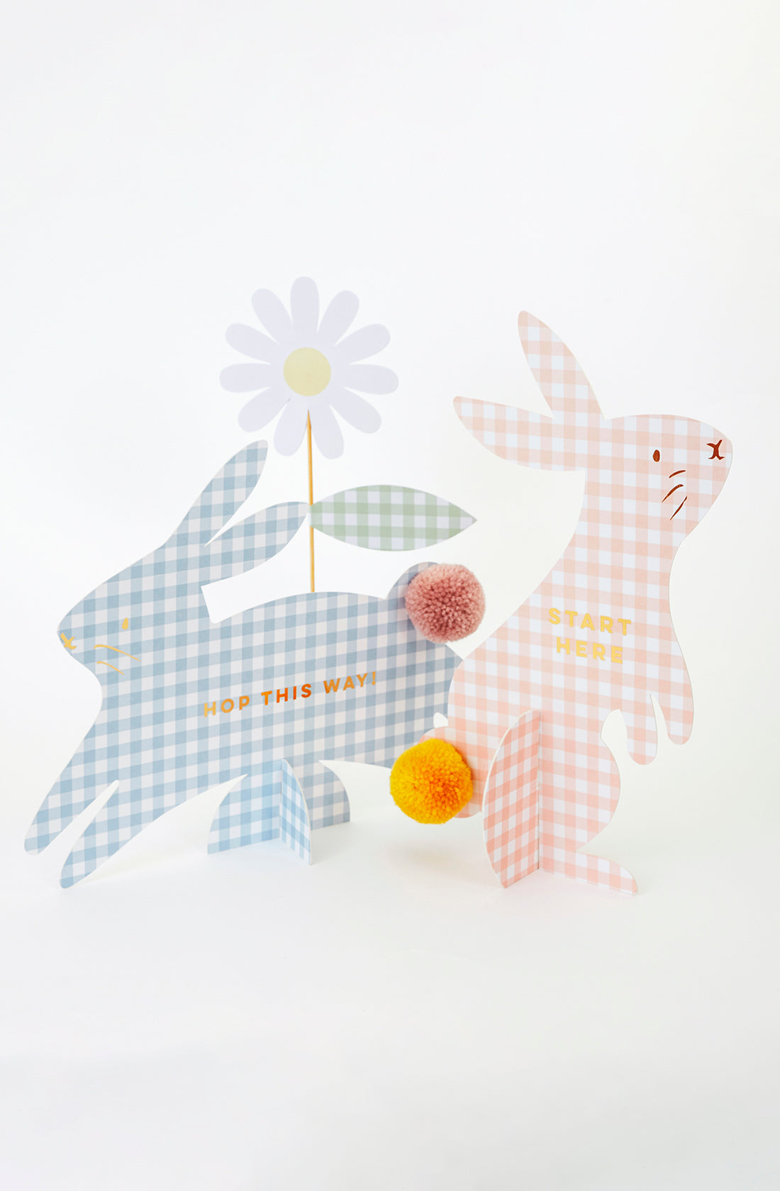 The Gingham Bunnies Egg Hunt Kit by MERI MERI includes two pastel cardboard gingham bunnies. The blue bunny says "Hop This Way" with a daisy, and the pink one reads "Start Here." Both have pom-pom tails, ideal for an egg hunt!.