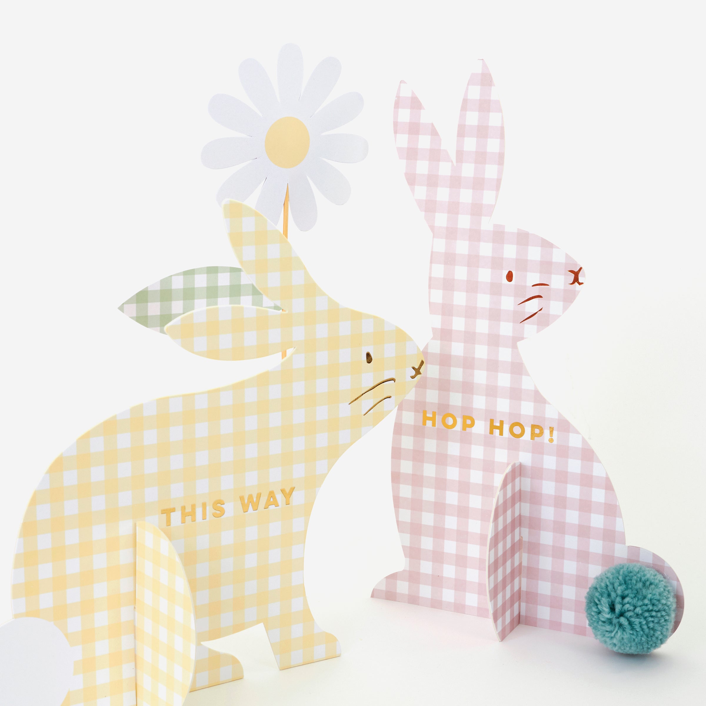 The Gingham Bunnies Egg Hunt Kit by MERI MERI features two charming paper bunnies with gingham prints: a yellow one saying "THIS WAY" with a pom-pom tail and a pink one declaring "HOP HOP!" complemented by a white daisy. This kit is perfect for an egg hunt, featuring elegant gold foil details.