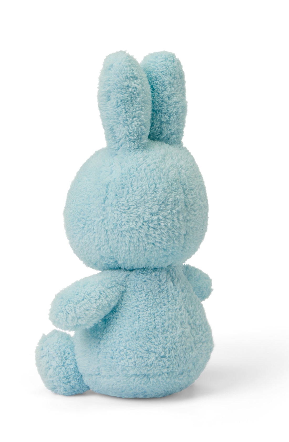 A plush toy from the BON TON TOYS X MIFFY Miffy Terry Collection, named Miffy Sitting Terry Light Blue 23 cm, is pictured facing away from the camera. The light blue bunny features a fluffy, textured appearance with long ears and a small tail and is filled with recycled polyester. The backdrop is plain white, making this adorable accessory an ideal baby gift!