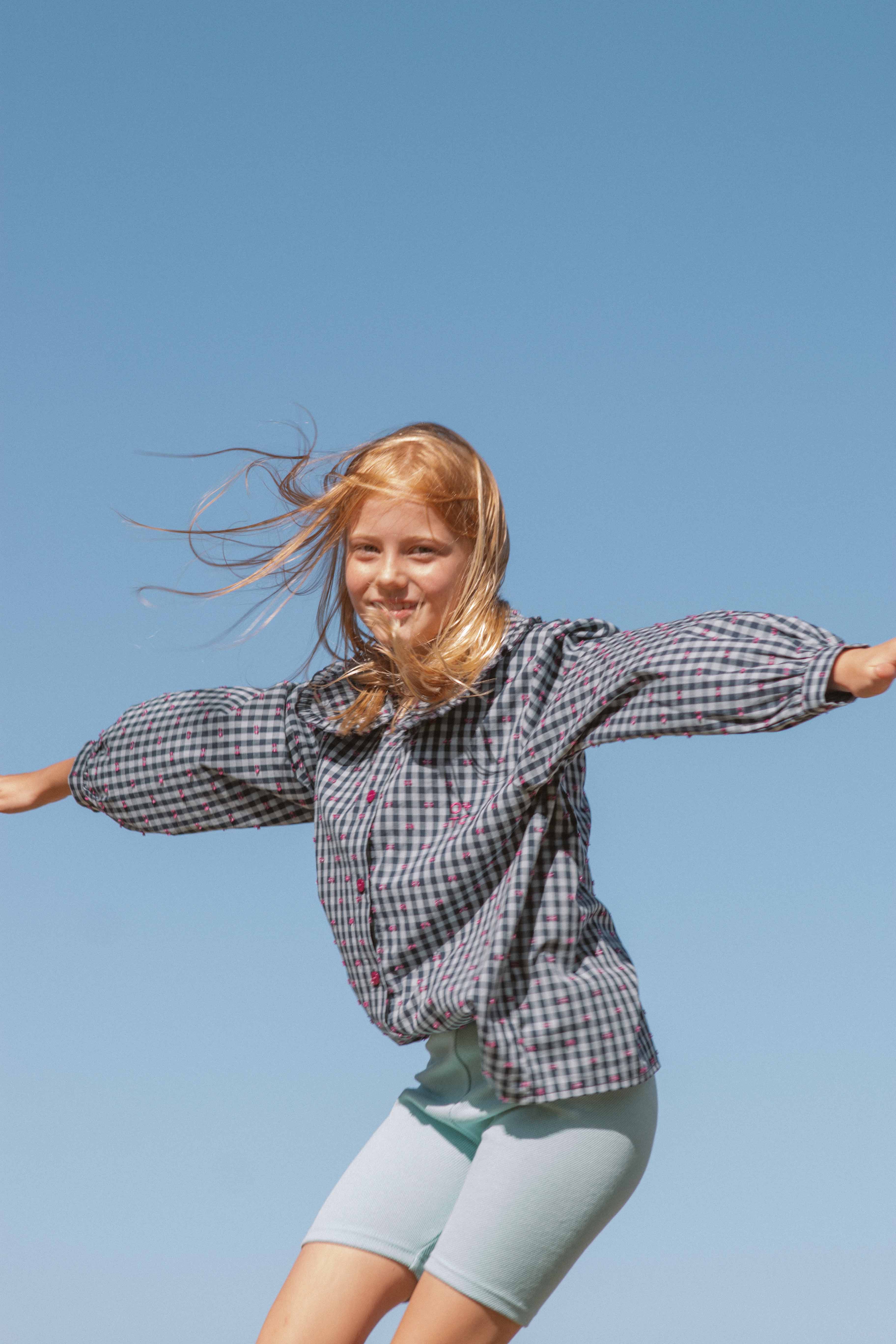 A young blonde girl, wearing a checkered shirt and Aqua Sky Bike Shorts by OLIVE + THE CAPTAIN, spreads her arms out against a clear blue sky. Her hair blows in the wind, and she has a joyful expression on her face.