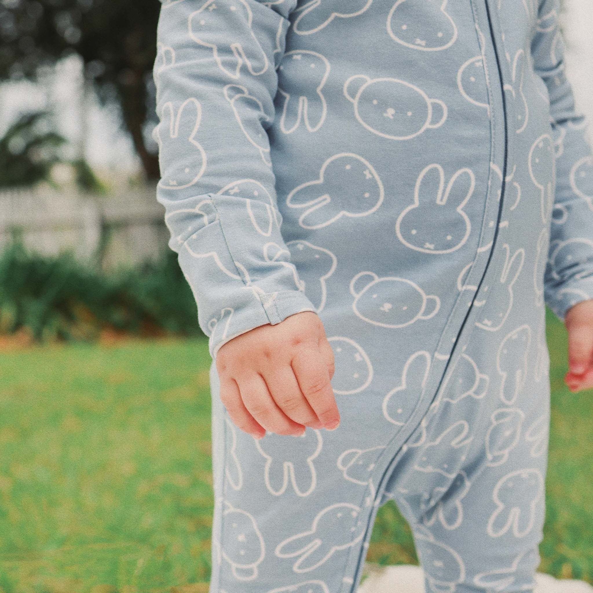 A child stands on green grass wearing the KYND BABY Miffy Day Or Night Onesie in Moody Blue, with only their lower face, body, and hand visible.