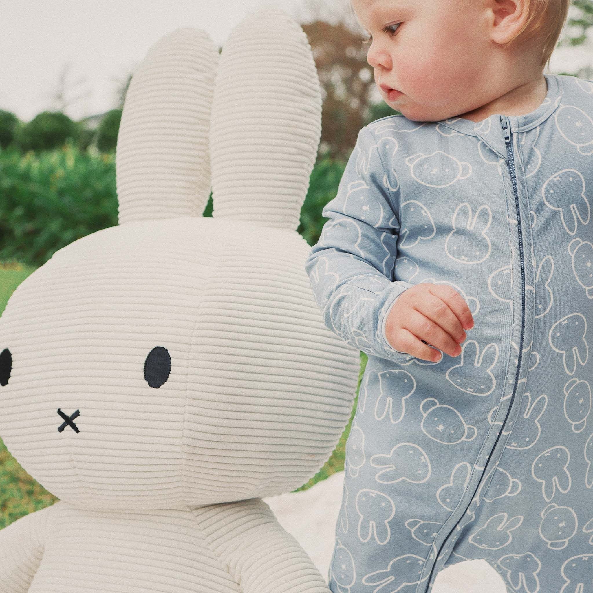 A baby wears the KYND BABY Kynd Baby - Miffy Day Or Night Onesie Moody Blue and stands outdoors on grass beside a large white corduroy stuffed bunny.