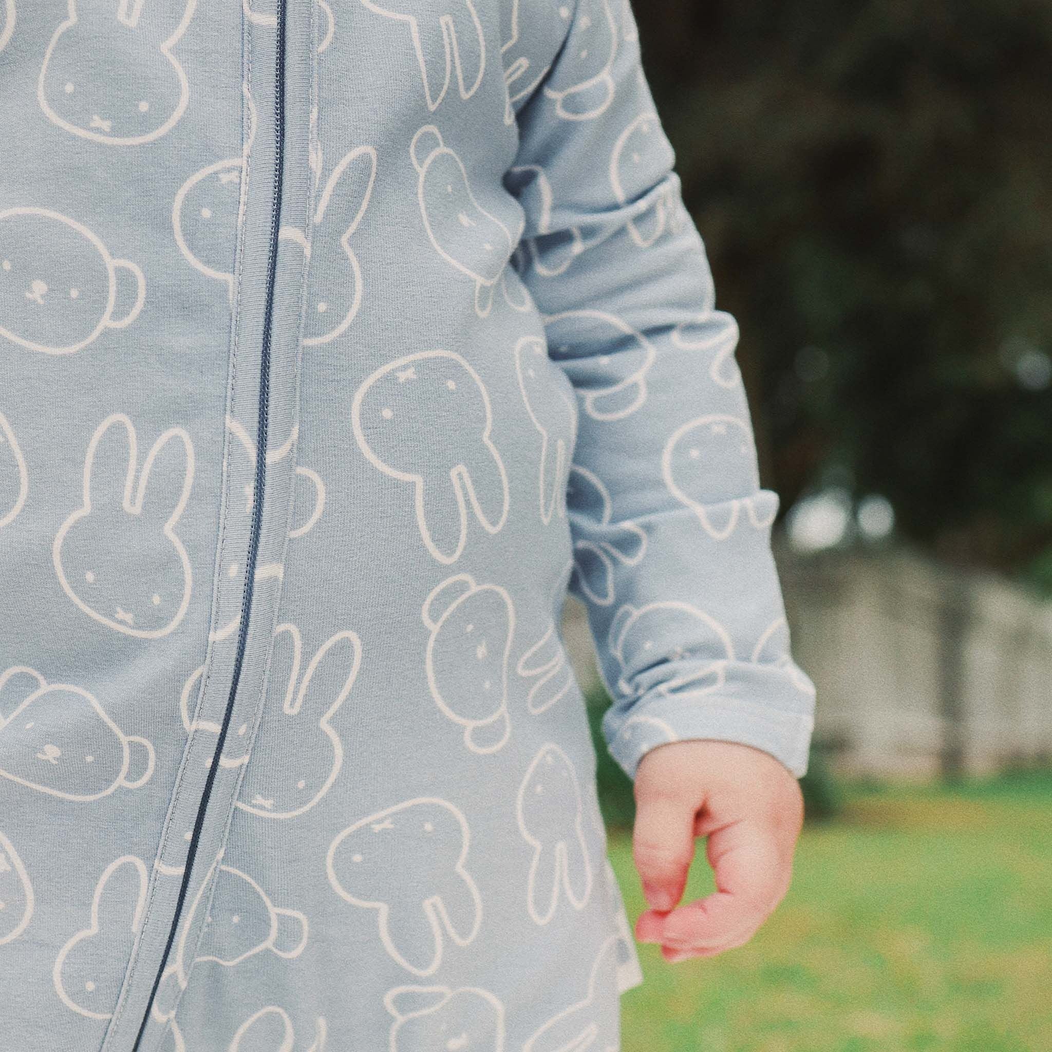 A child wears the KYND BABY Miffy Day Or Night Onesie in Moody Blue, featuring white bunny faces, while standing outdoors on grass with trees and a fence softly blurred in the background.
