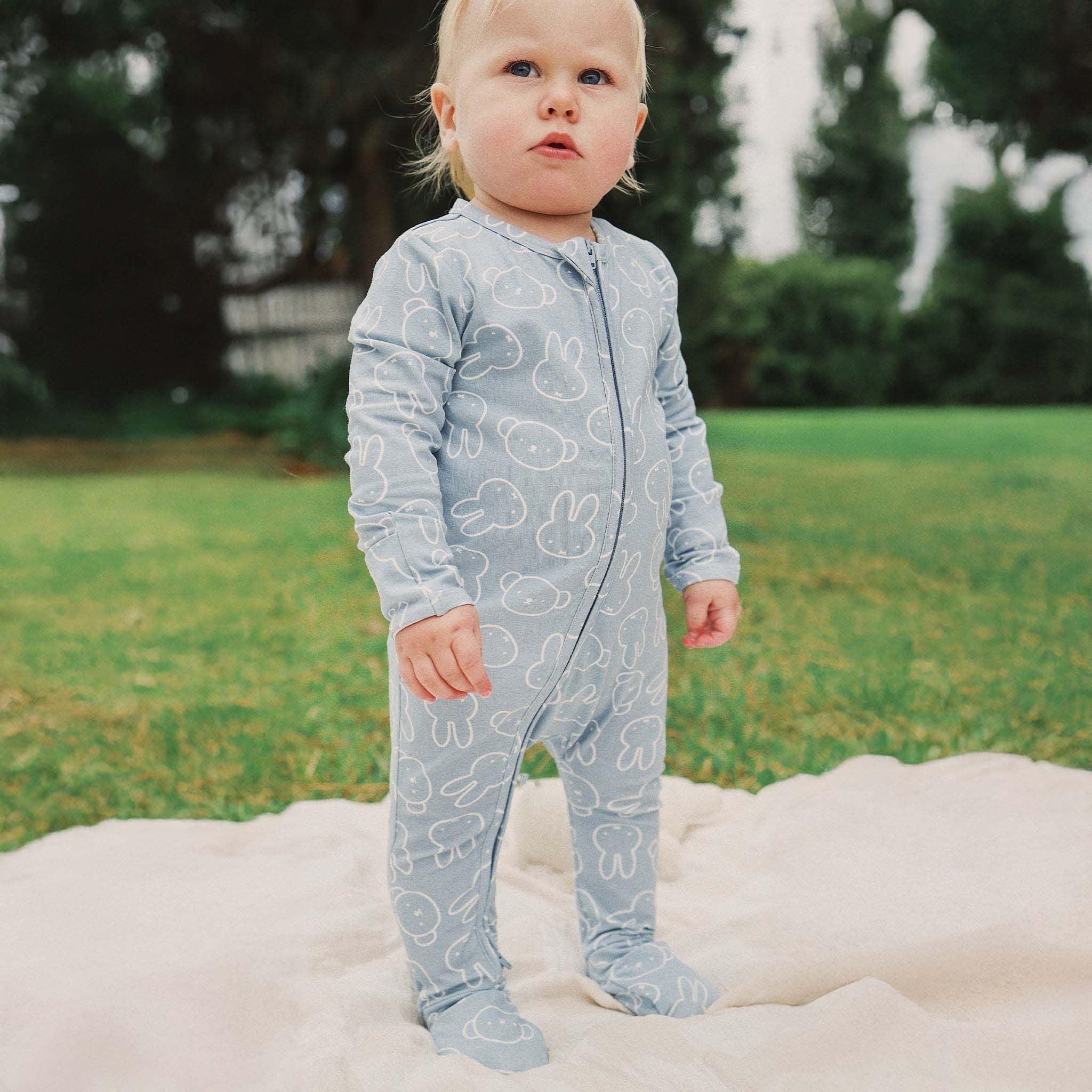 A toddler wearing the KYND BABY Kynd Baby - Miffy Day Or Night Onesie Moody Blue with white bunny patterns stands on a light blanket outdoors, surrounded by grass and trees.