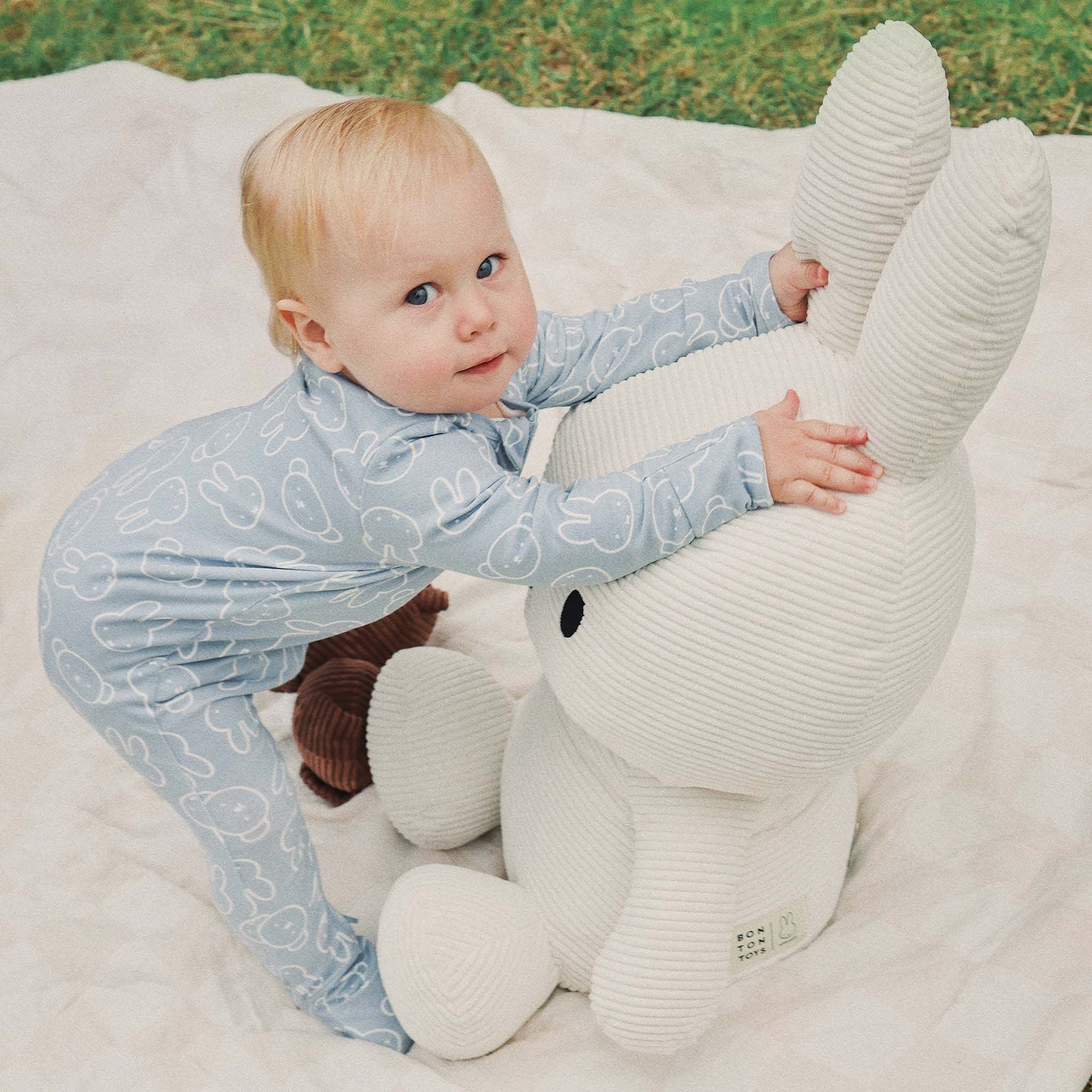 A baby wearing the KYND BABY Miffy Day Or Night Onesie in Moody Blue stands on a blanket, holding a large white plush bunny, on a grassy area.