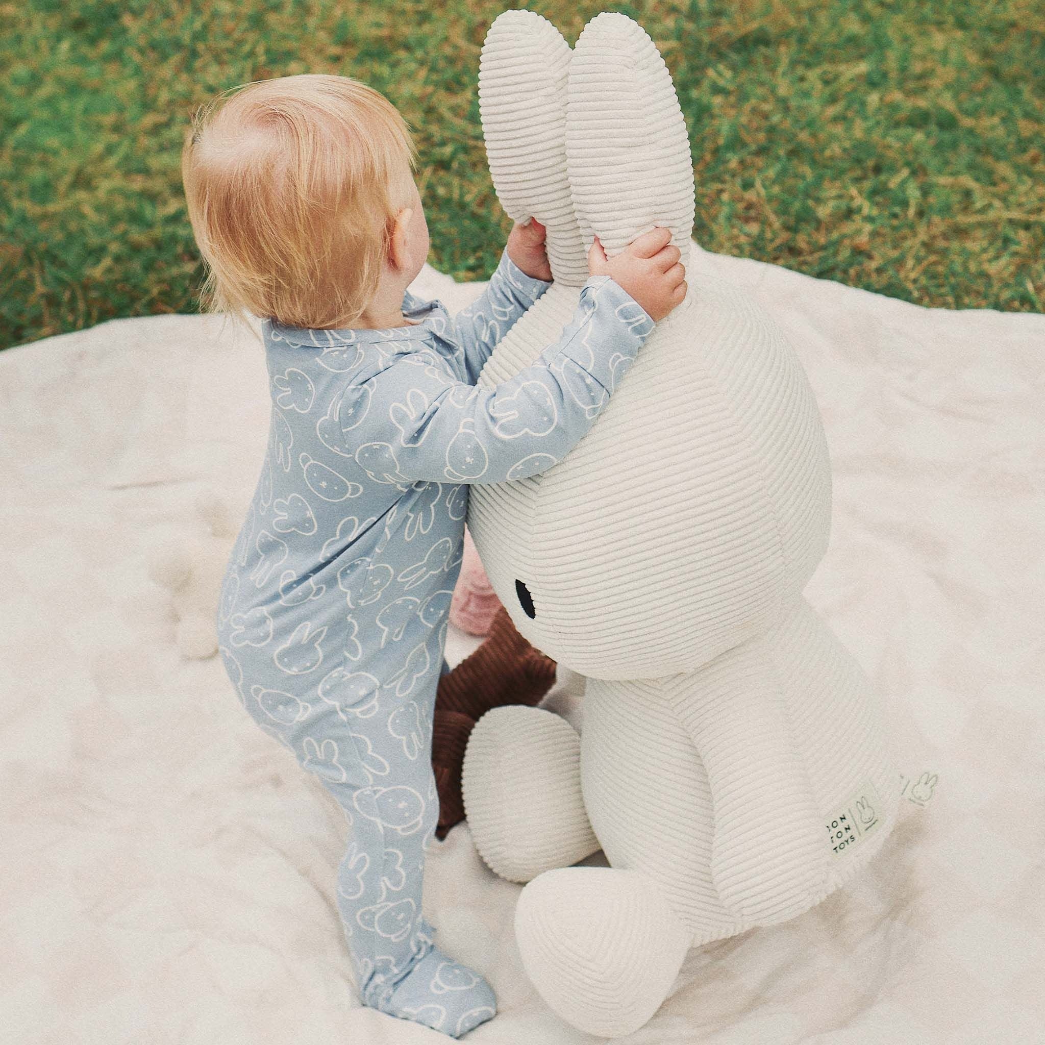 A toddler in the KYND BABY - Kynd Baby Miffy Day Or Night Onesie Moody Blue stands on a blanket outdoors, touching the ears of a large white stuffed bunny toy, with grass visible in the background.