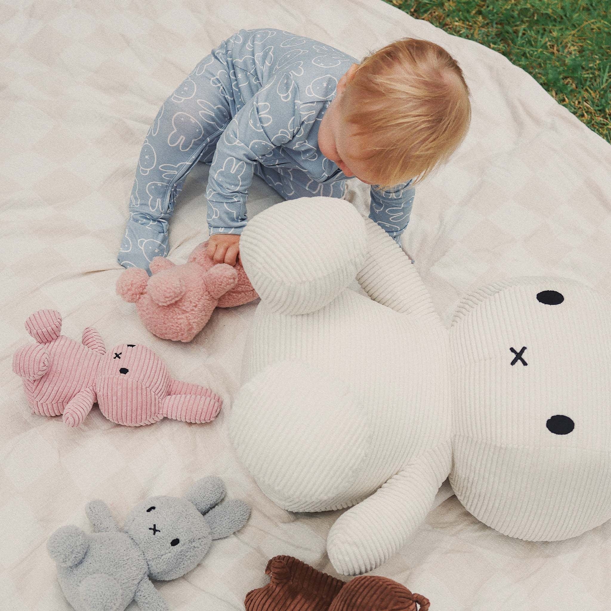 A baby wears the KYND BABY Miffy Day Or Night Onesie in Moody Blue, sitting on a blanket on the grass and reaching for white, pink, gray, and brown stuffed bunny toys.