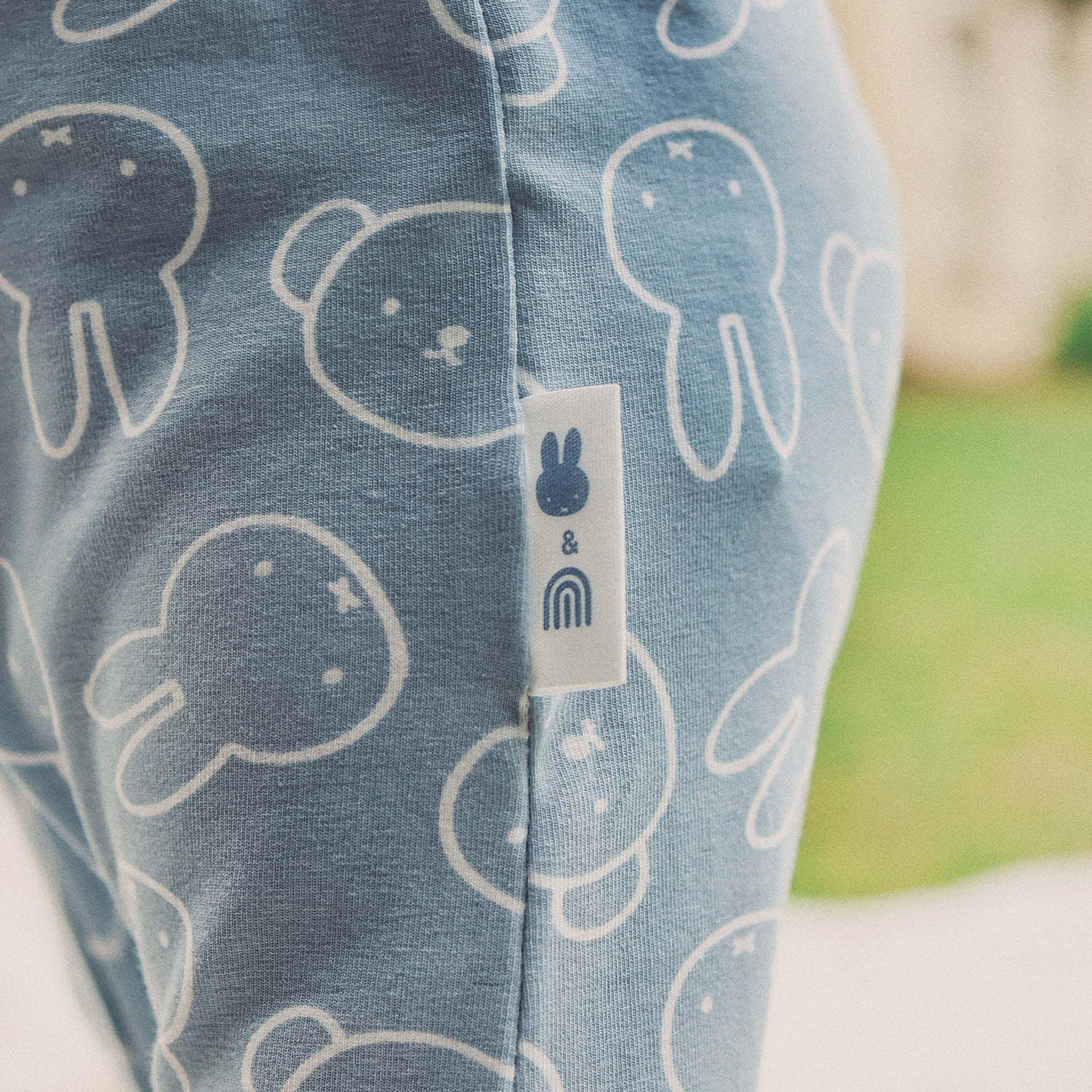 Close-up of the KYND BABY Kynd Baby - Miffy Day Or Night Onesie in Moody Blue, featuring white bunny line drawings and a rainbow bunny tag on soft organic cotton, shown against a blurred green and white background.