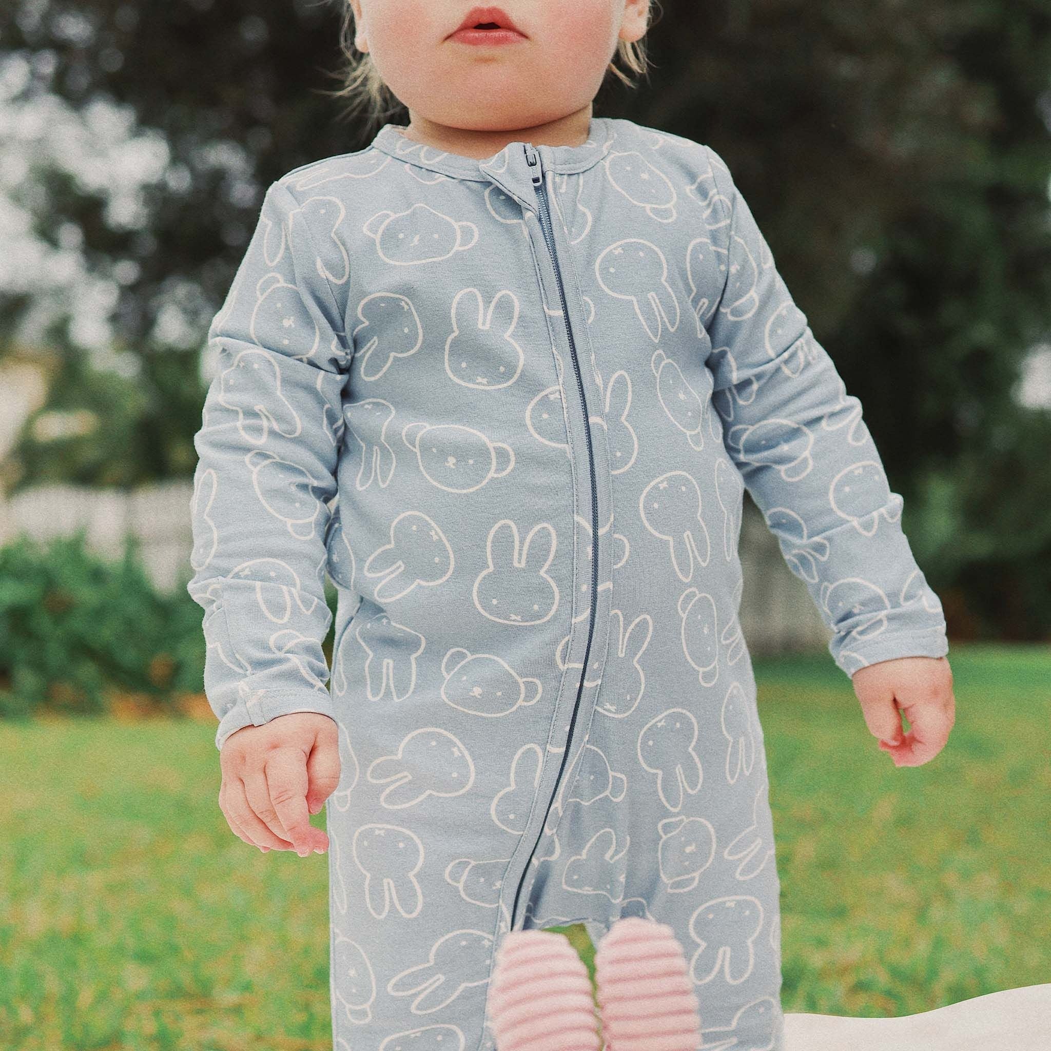 A young child in the KYND BABY "Kynd Baby - Miffy Day Or Night Onesie Moody Blue" stands on grass outdoors, with white bunny designs and greenery blurred in the background. The child's face is partially visible.