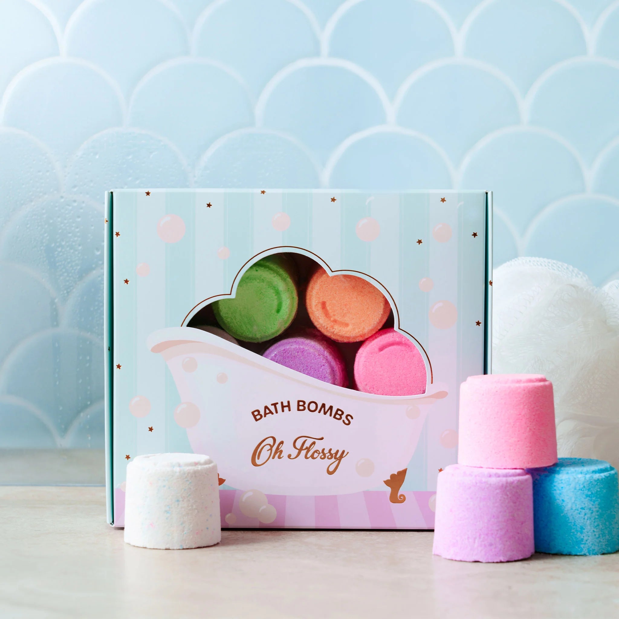 A box of Oh Flossy ~ Kids Mini Bath Bombs, featuring four colorful, handmade-in-Australia treasures by OH FLOSSY, is displayed. Pink, purple, and white mini bath bombs rest in front of the box against a soft blue scallop-patterned wall backdrop. Gentle on kids' skin.