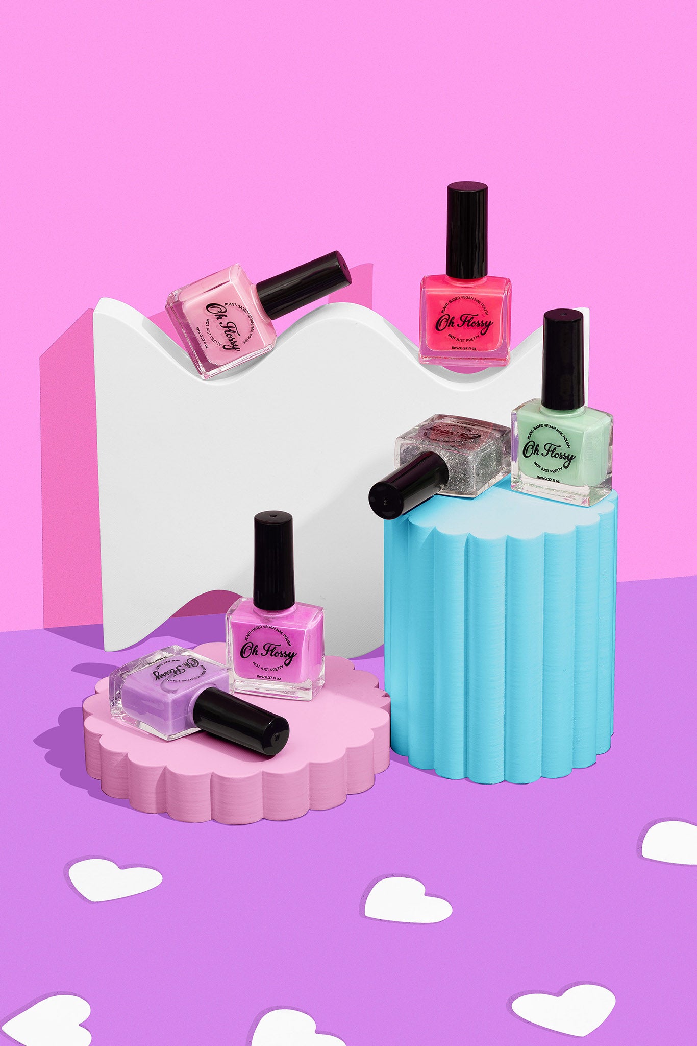 Five OH FLOSSY Oh Flossy - Nail Polish Pastel Pink bottles are displayed on pastel pink and blue platforms, set against a pink and purple backdrop with white heart-shaped cutouts scattered around.