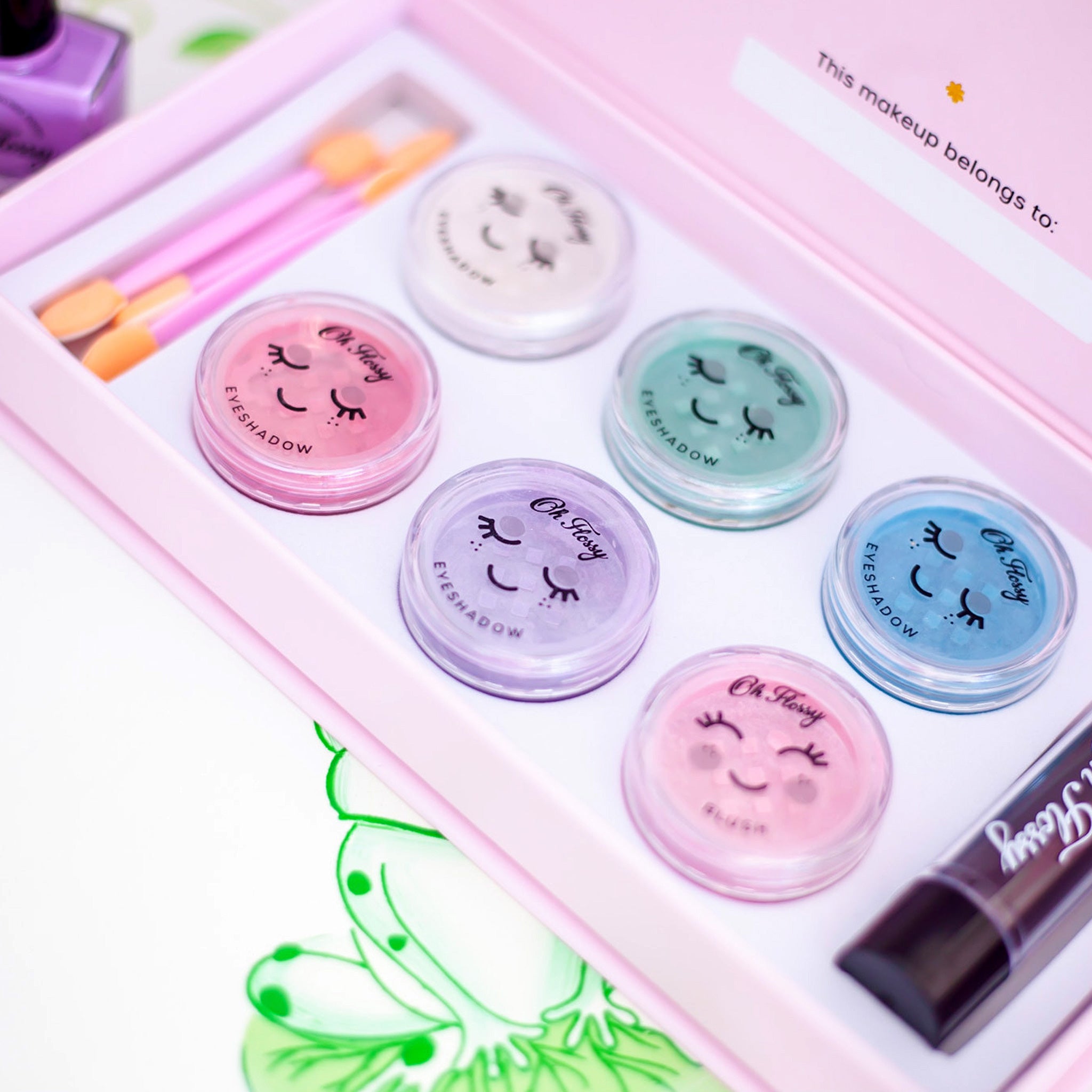Unbox the Oh Flossy ~ Deluxe Makeup Set by OH FLOSSY, featuring six pastel, non-toxic eyeshadow containers with cheerful faces. Includes brushes and a makeup tube. The box lid has a "This makeup belongs to" label—ideal for kids discovering natural cosmetics!.