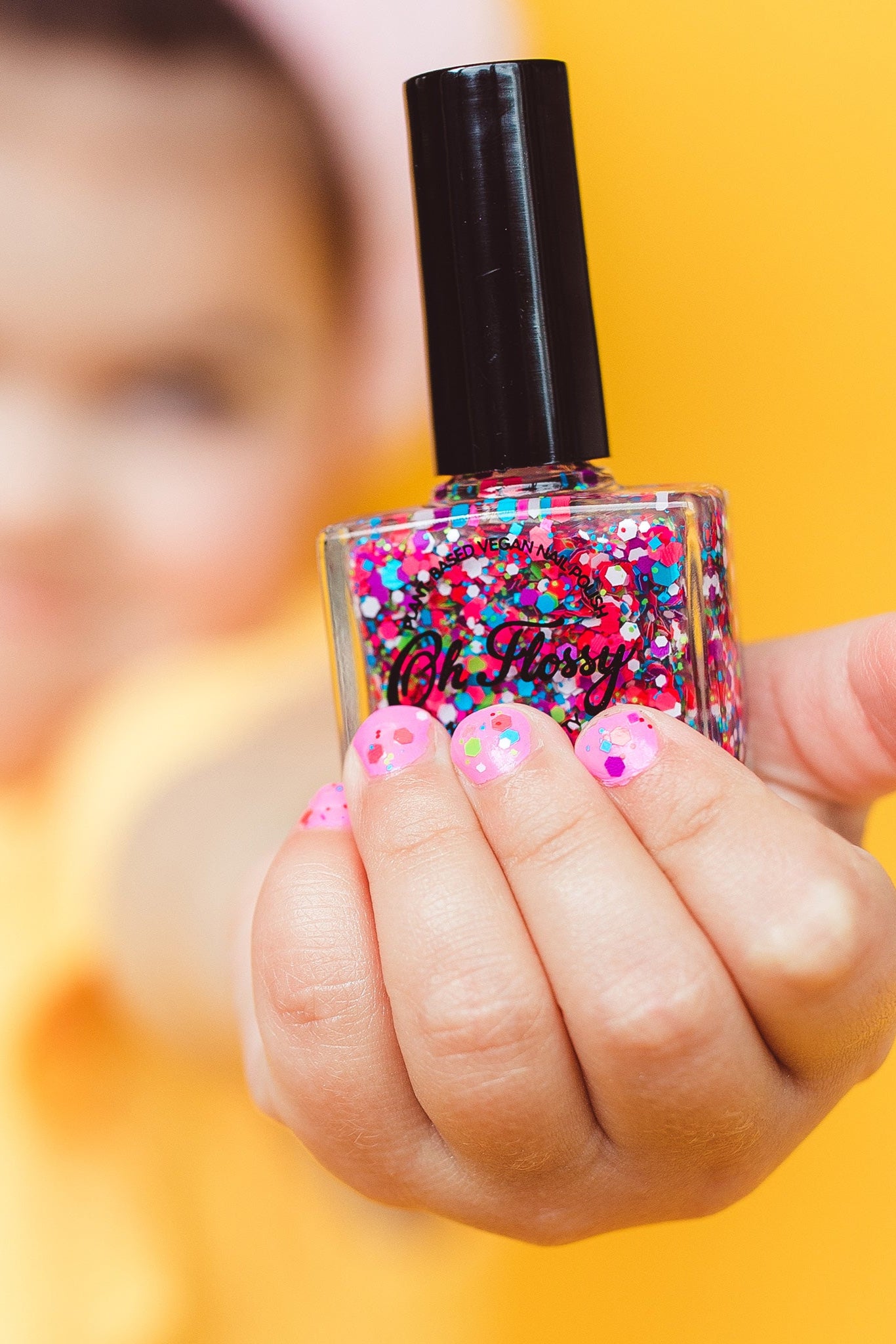 Close-up of a hand with nails painted in glittery pink from the Oh Flossy ~ Party Nail Polish Set by OH FLOSSY, holding the vegan-friendly bottle. The background is a blurred yellow, complementing the vibrant pink and colorful glitter on the nails.