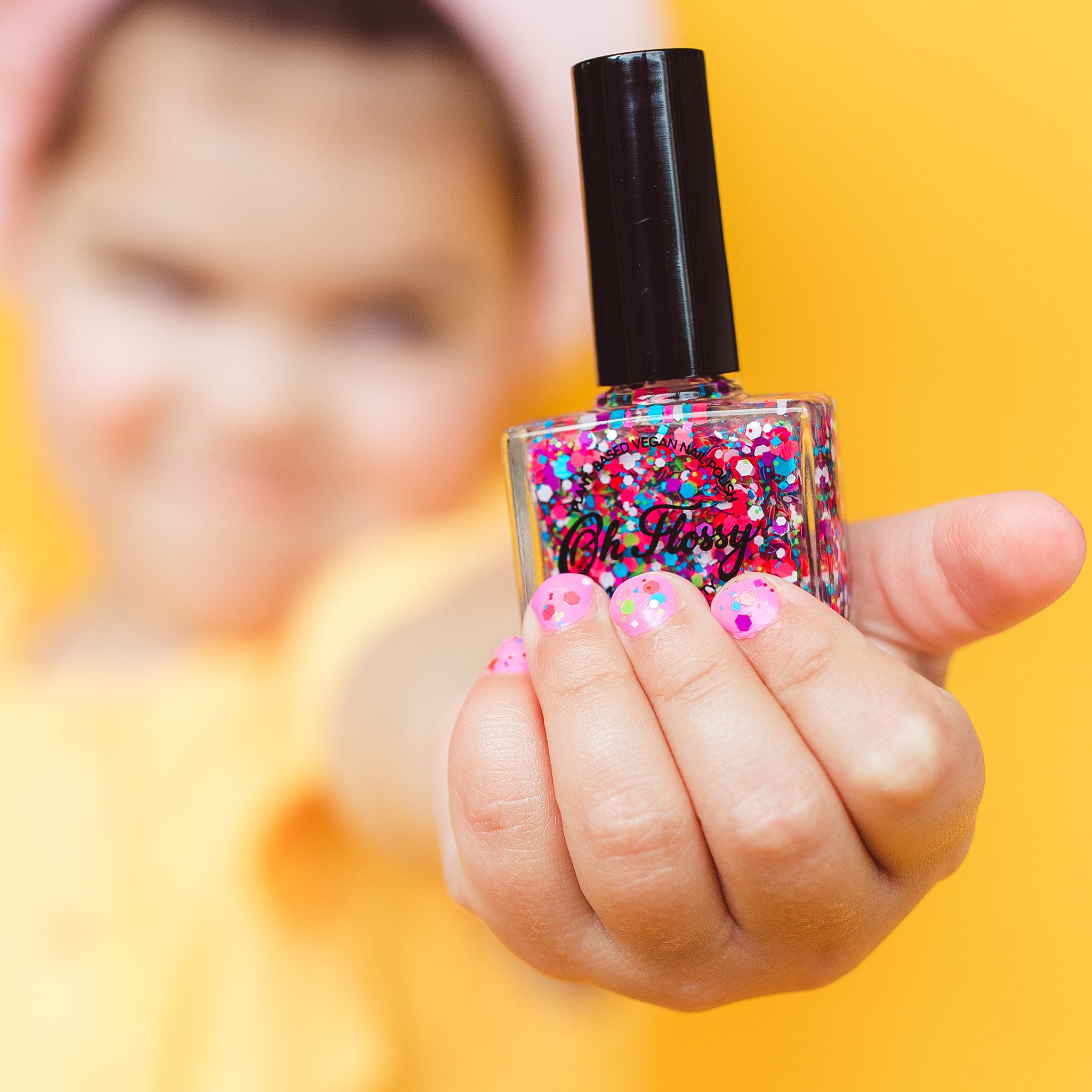 A person holds a bottle of Oh Flossy ~ Nail Polish Confetti Glitter by OH FLOSSY, showcasing their nails adorned with pink and blue sparkles. The sunny yellow backdrop accentuates the lively hues and plant-based, 10-Free formula, ideal for kids' nail polish adventures.