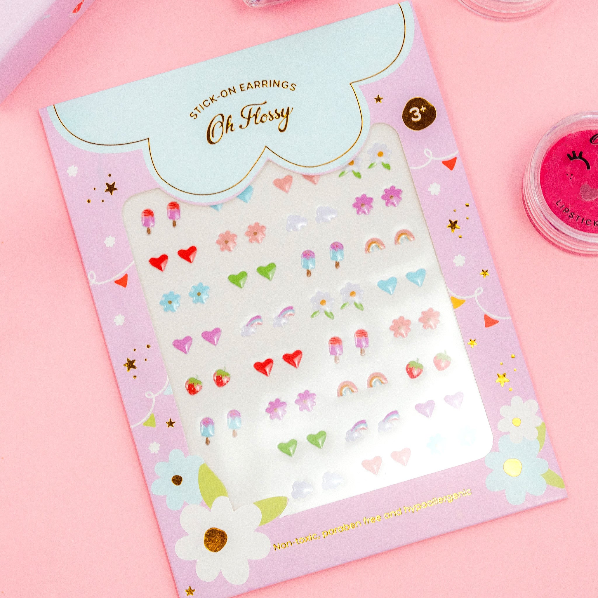 The "Oh Flossy ~ Kids Stick-On Earrings" by OH FLOSSY are vibrant, non-toxic accessories for kids. Displayed on a decorative card, the earrings come in fun shapes like hearts, stars, and rainbows. The packaging has playful designs on a pink backdrop—ideal for charming children's accessories.