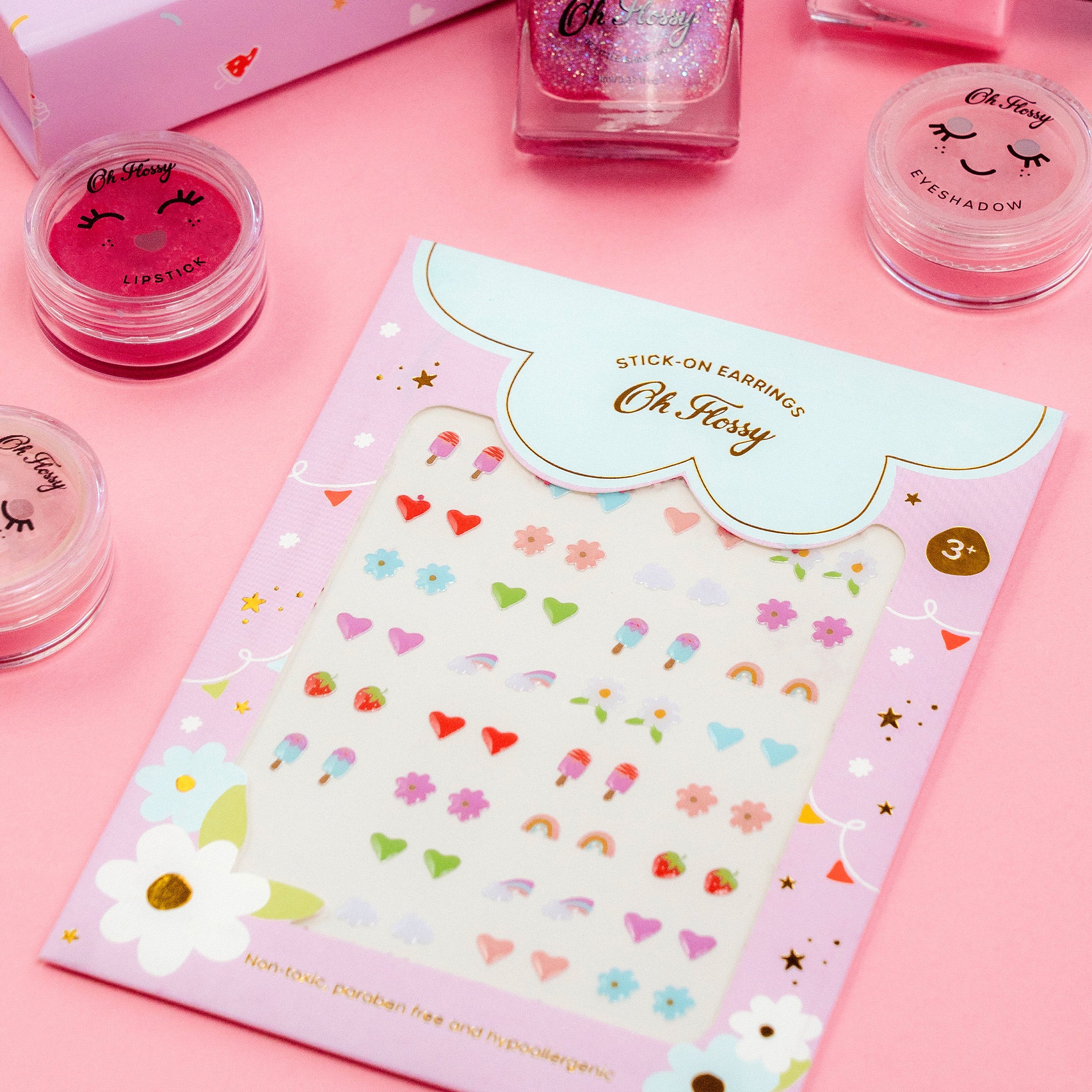 A packet of Oh Flossy ~ Kids Stick-On Earrings is set on a pink surface, made from non-toxic materials with playful heart, flower, and rainbow shapes. Nearby are pink and glittery makeup jars labeled lipstick and eyeshadow.