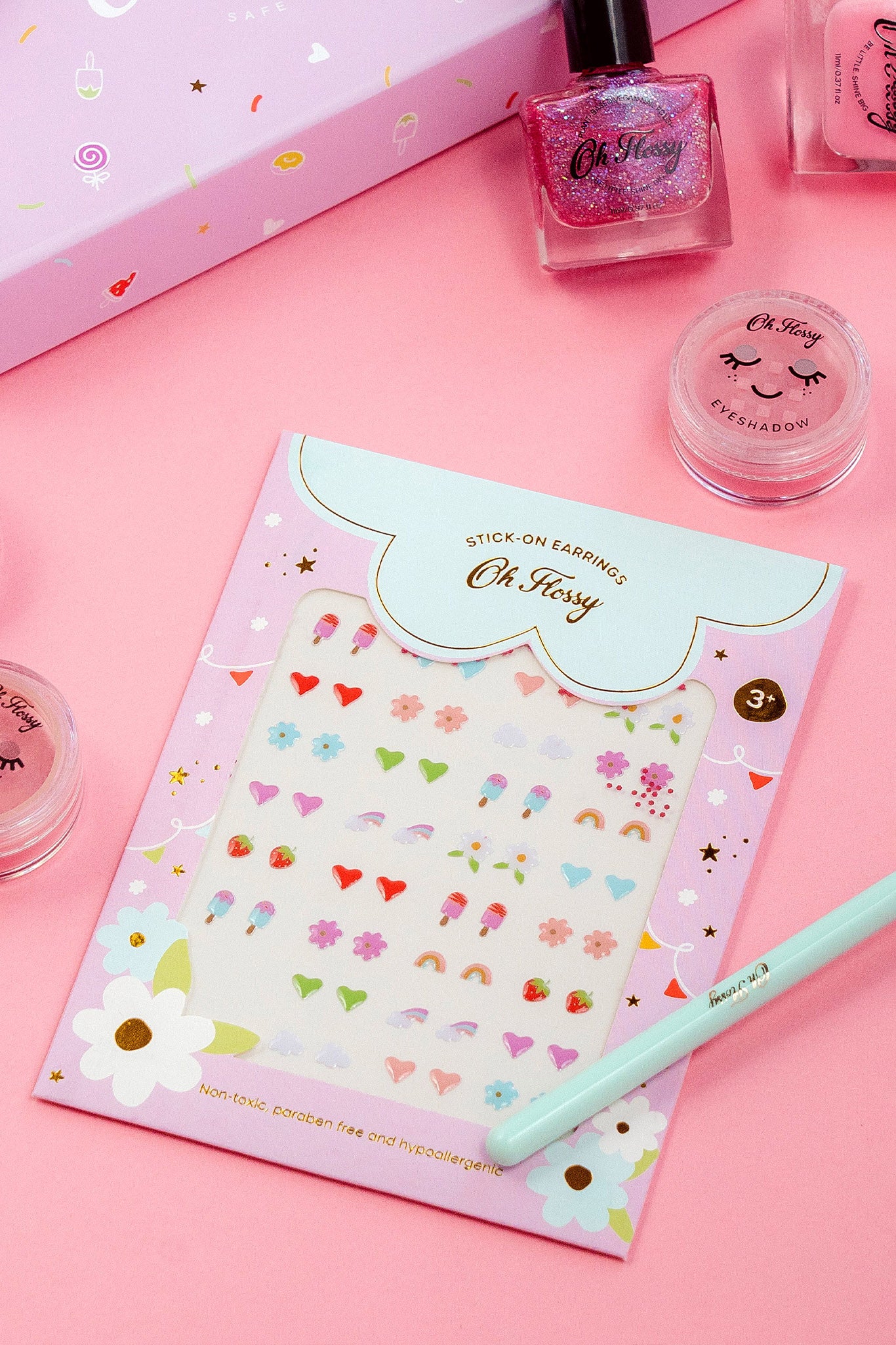 Oh Flossy ~ Kids Stick-On Earrings by OH FLOSSY are colorful and non-toxic, featuring hearts, flowers, and rainbows. Displayed on an "Oh K!" card alongside pink nail polish and cosmetics on a pink background, these charming accessories make delightful additions to any collection.