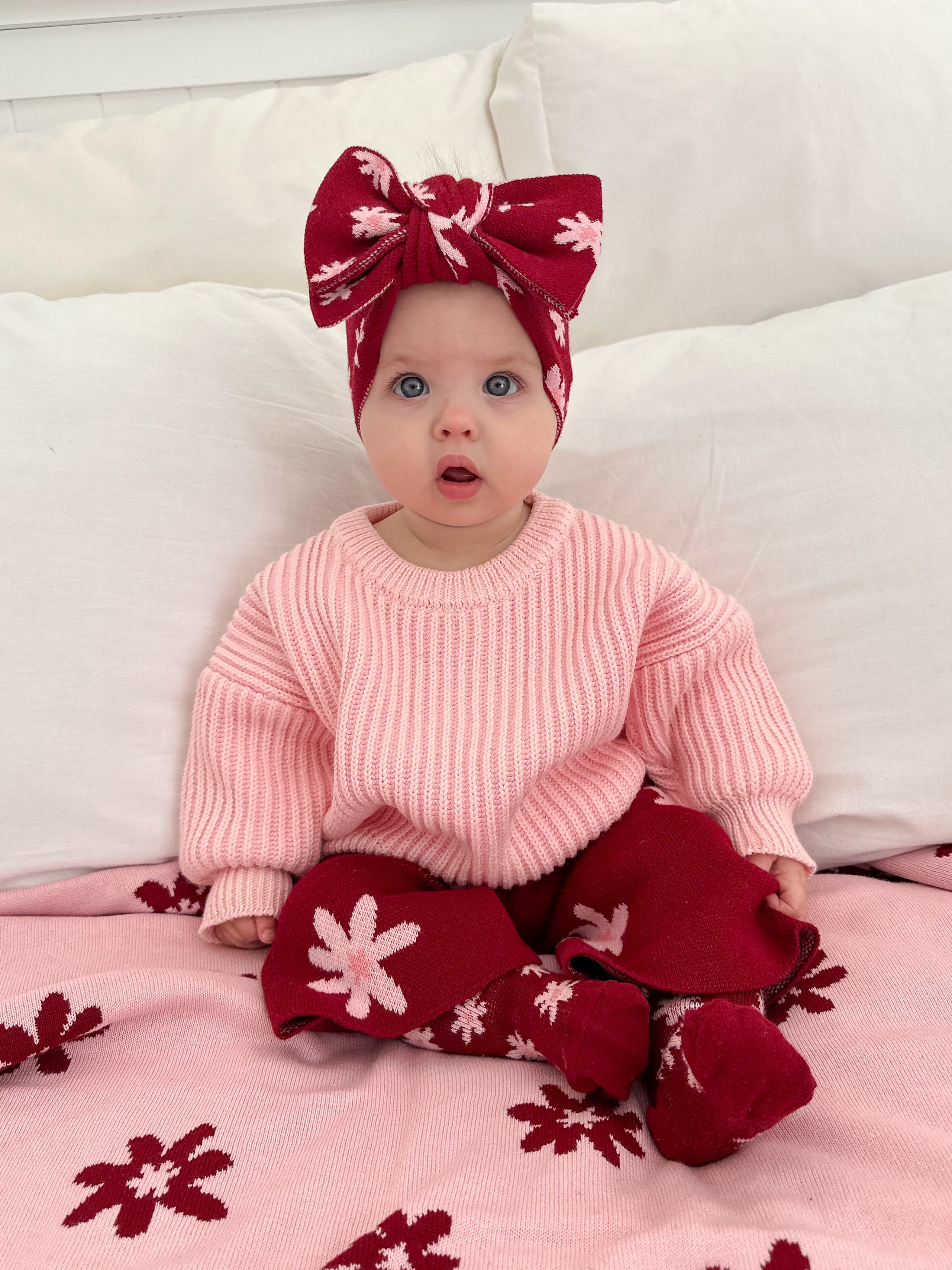 A baby in a pink sweater, red leggings, pink flower socks, and the ZIGGY LOU Ziggy Lou - Headband Lola (Plum) with a large bow sits on floral blankets, looking up with a surprised expression.