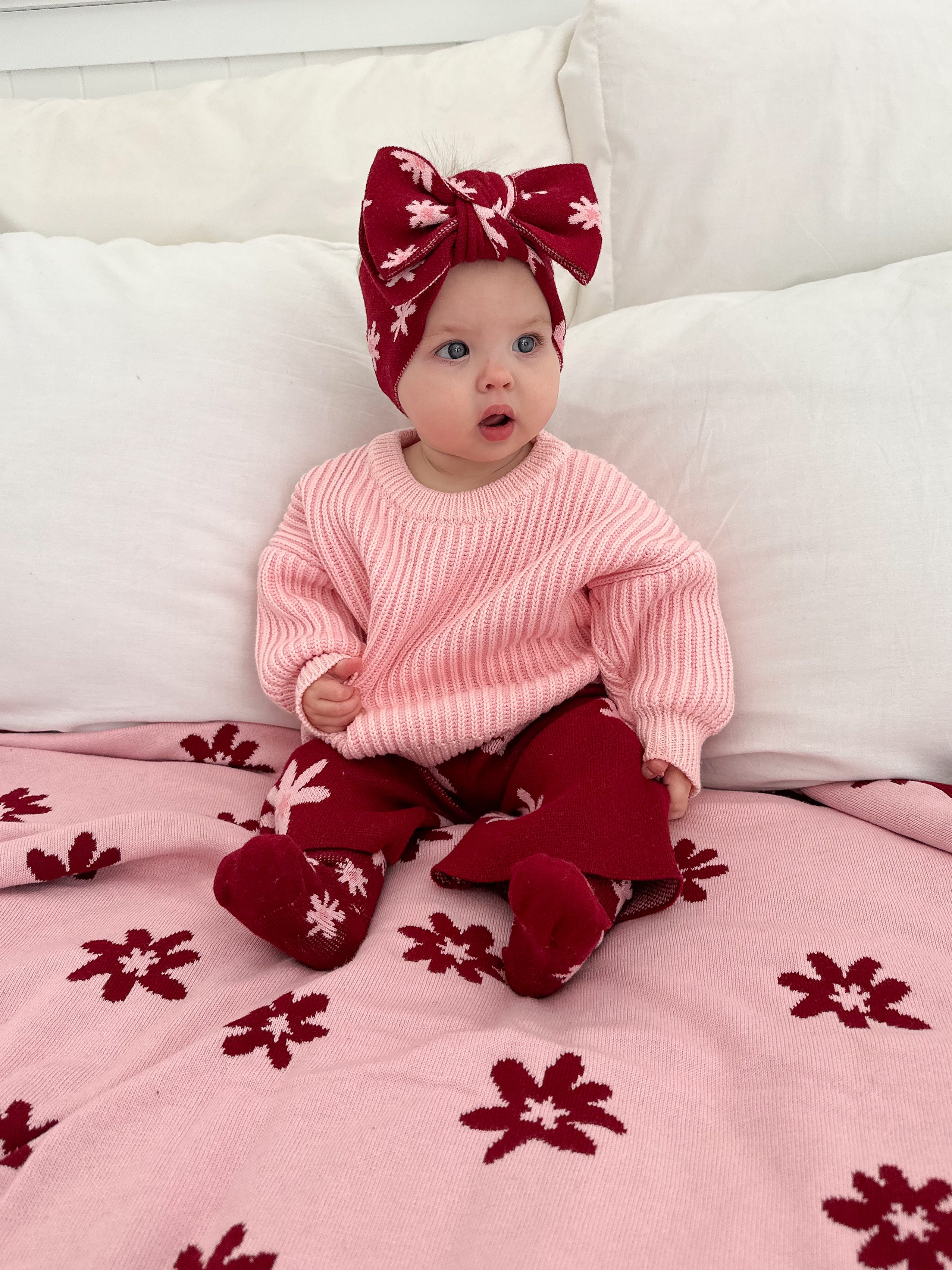 A baby wearing a large ZIGGY LOU Ziggy Lou - Headband Lola (Plum), a pink sweater, red pants, and matching socks sits on a bed with a pink and red floral blanket, looking slightly to the side with an open-mouthed expression.