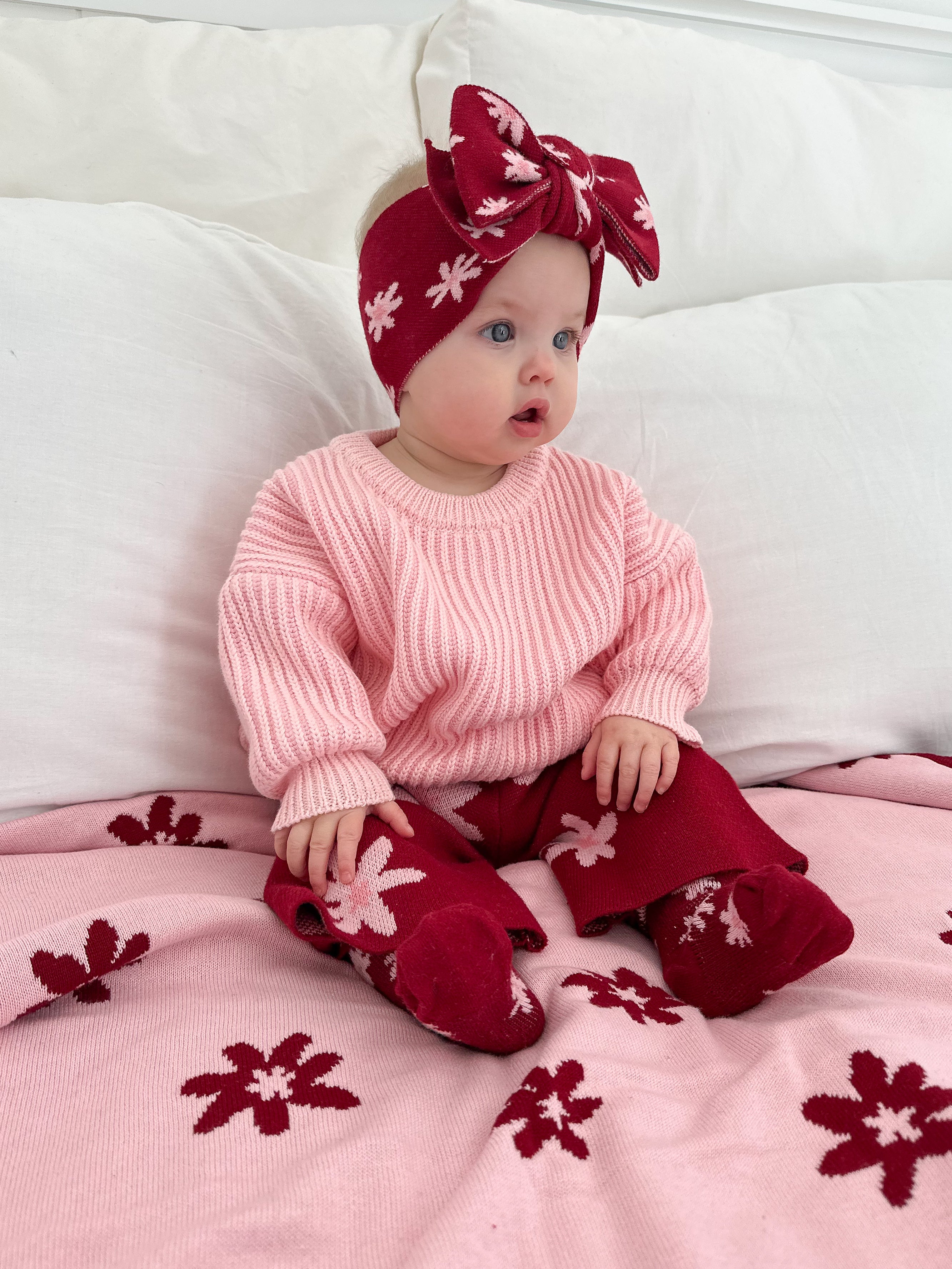 A baby wearing a pink sweater, red floral pants, matching socks, and a ZIGGY LOU Ziggy Lou - Headband Lola (Plum) sits on a bed with a pink and red floral blanket.
