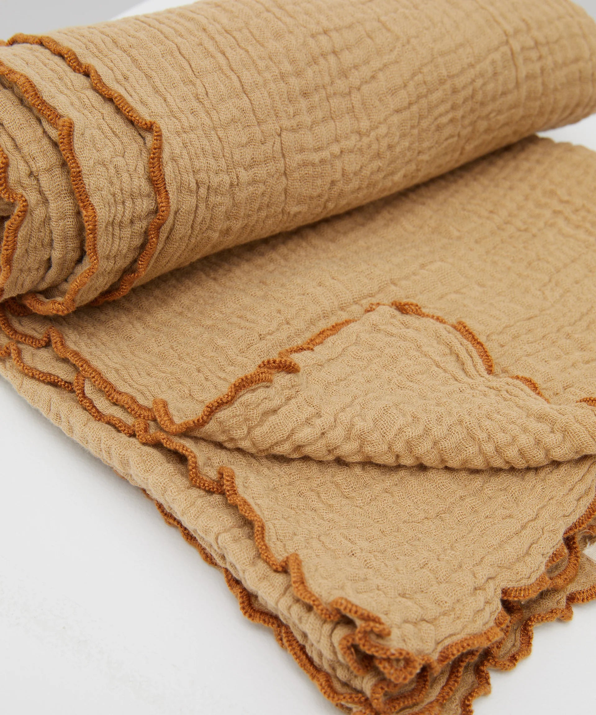 A close-up of the Philé - Swaddle Cacao by PHILÉ, a beige textured muslin wrap from the Cotton Classics Collection, crafted from Oeko-Tex Certified Cotton with wavy orange-stitched edges, shown folded against a white background.
