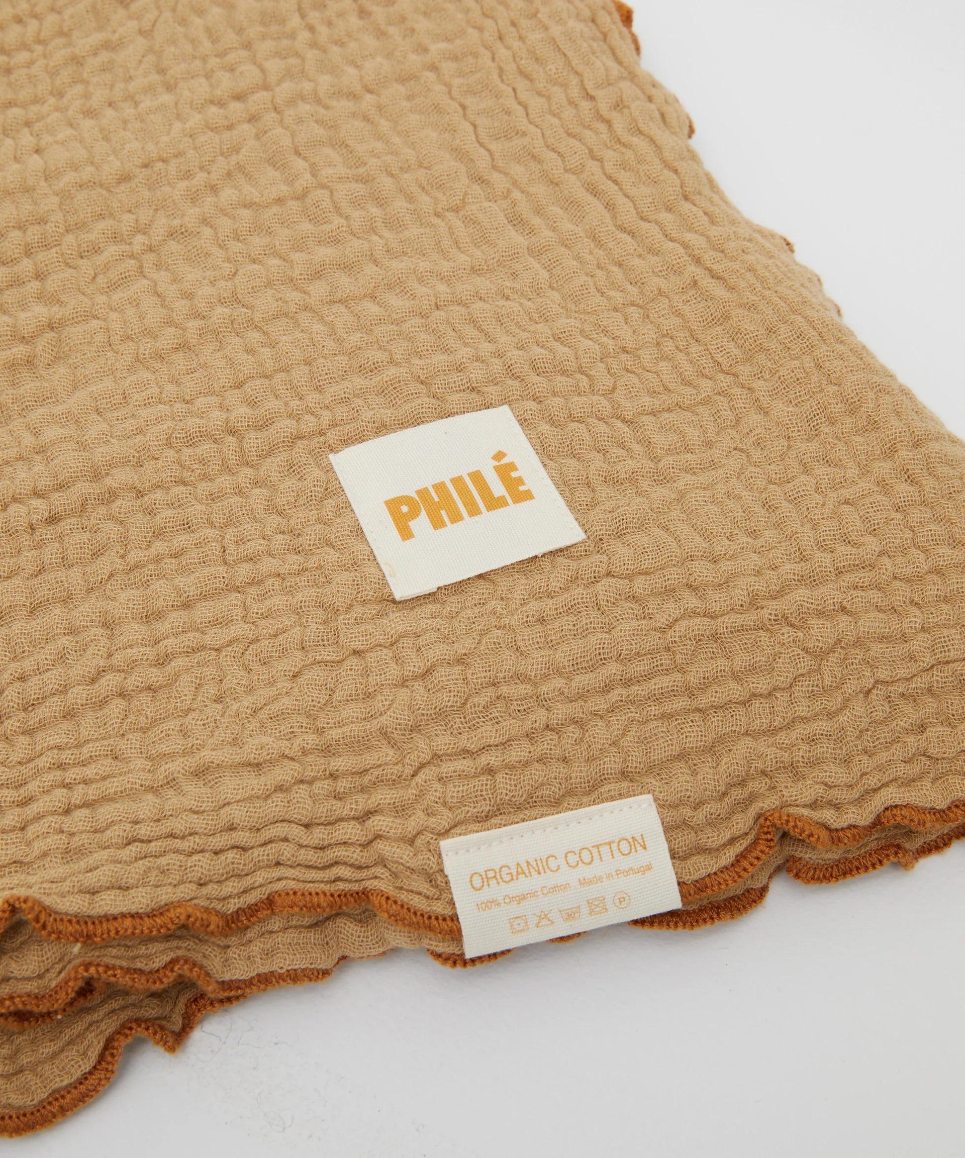 A close-up of the Philé - Swaddle Cacao by PHILÉ shows its textured beige fabric and two labels—"PHILÉ" and "ORGANIC COTTON 100% Organic Cotton, Made in Portugal," with care symbols. Oeko-Tex Certified for quality and safety.