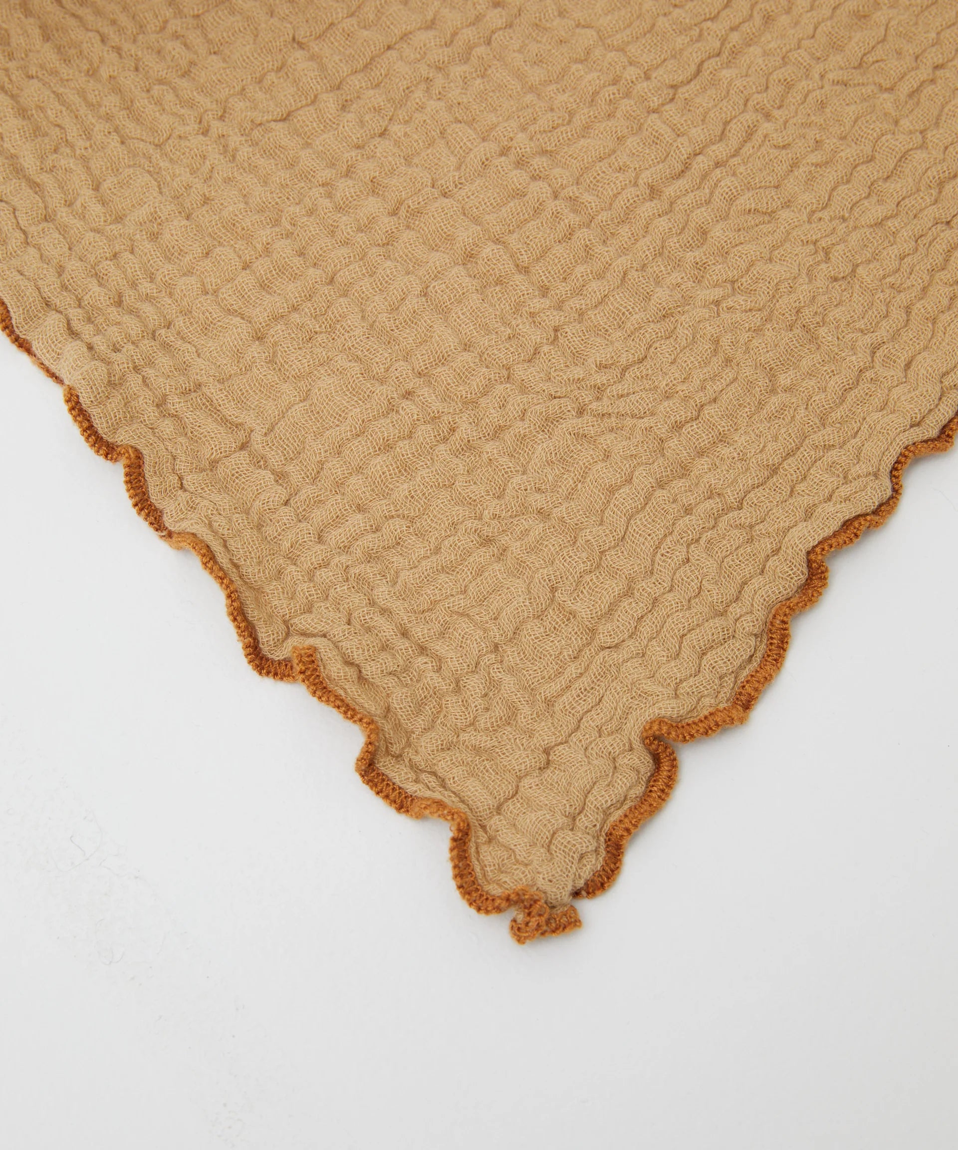 A close-up of the PHILÉ Philé - Swaddle Cacao, an Oeko-Tex certified cotton muslin wrap with beige textured fabric and an orange scalloped edge, laid flat on a white surface.