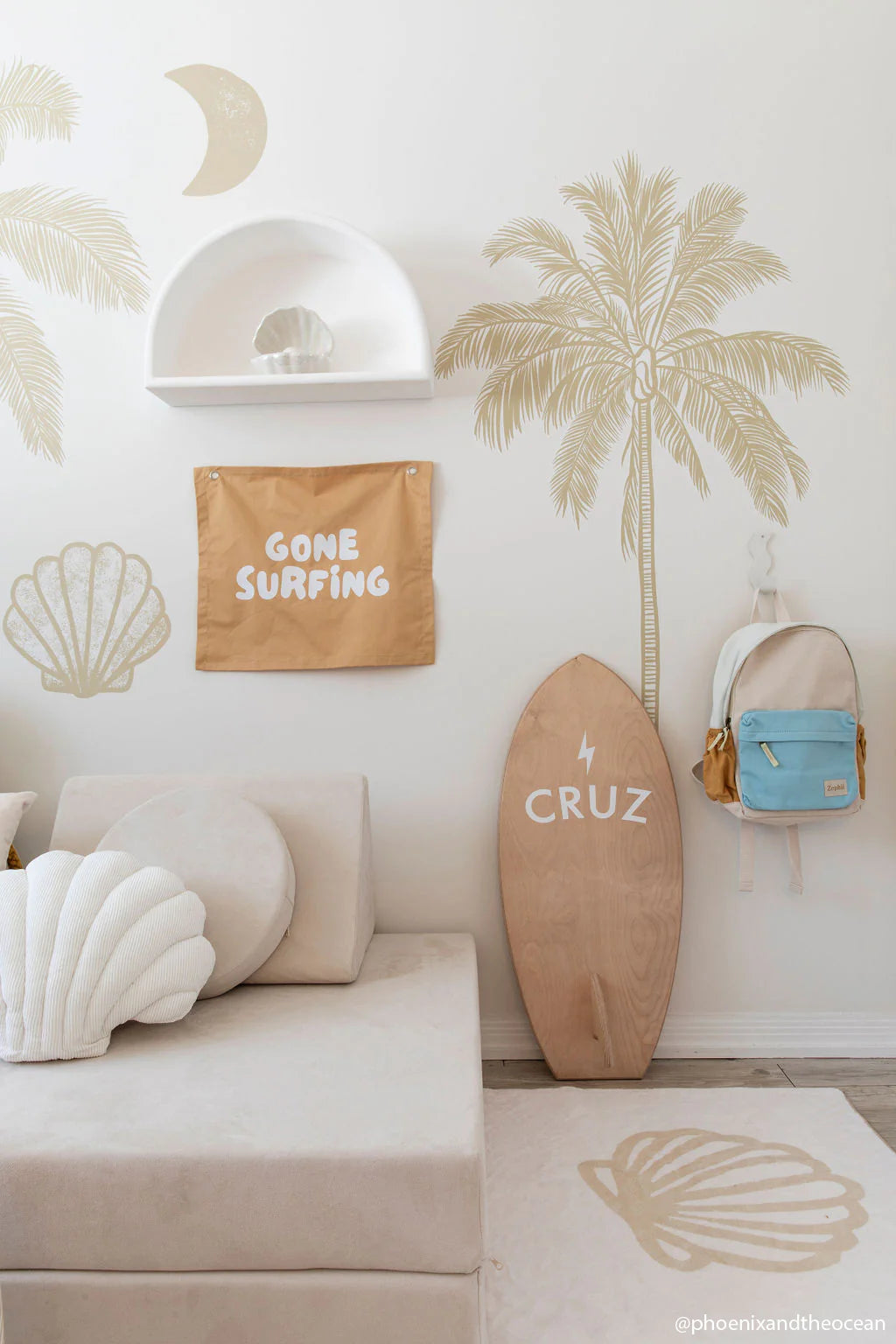 A cozy room with a beach theme highlights palm tree and shell illustrations on the wall, a "Gone Surfing" banner, and a surfboard with "CRUZ" next to it. Nearby hangs the ZEPHII Jagger Backpack featuring adjustable straps, while a white seashell pillow is placed on the light sofa.