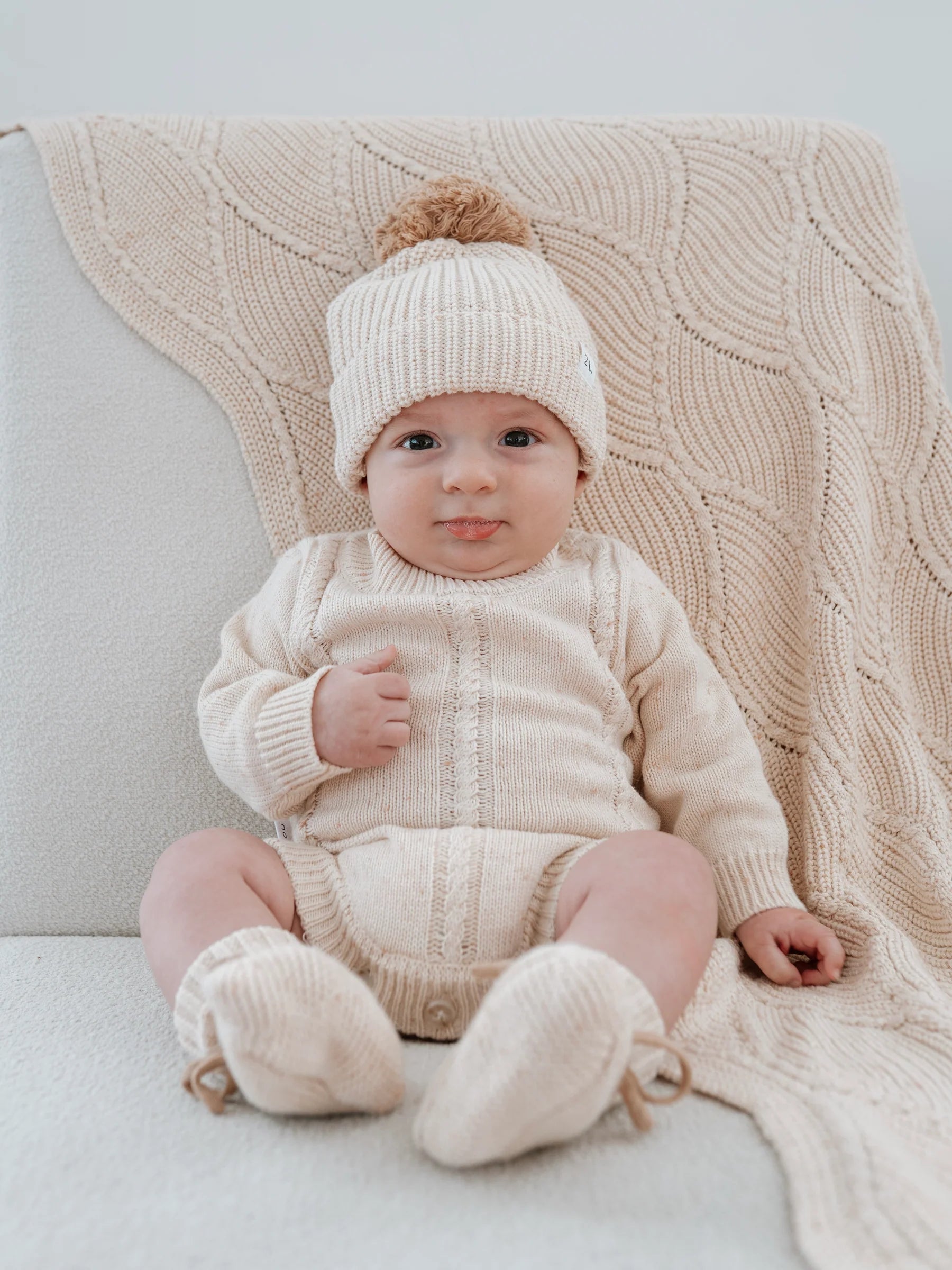 A baby sits on a cream-colored chair, smiling at the camera, dressed in a 100% cotton knitted outfit with hat and booties. The ZIGGY LOU Ziggy Lou - Blanket Biscotti Fleck is draped over the chair.