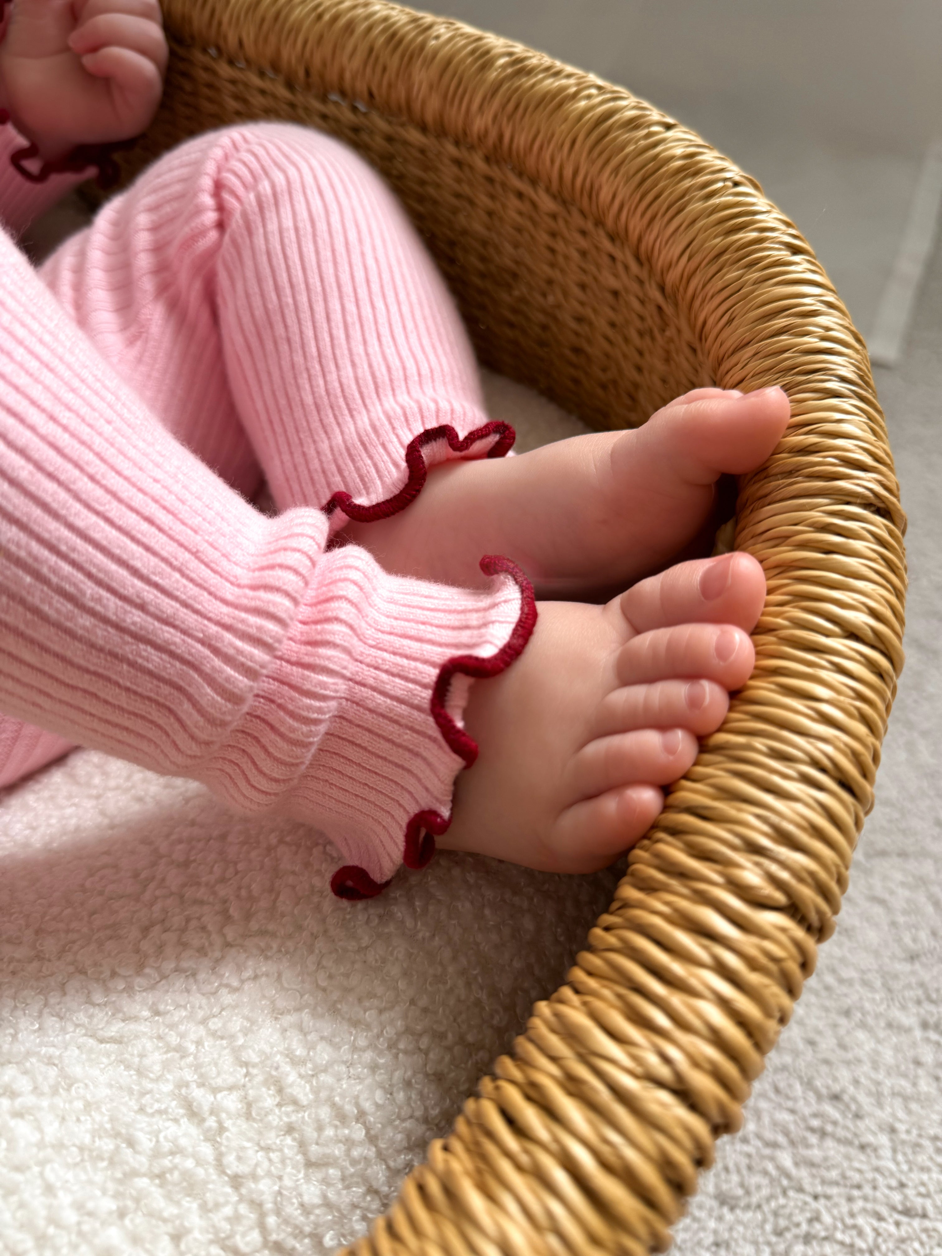 A close-up of a baby’s feet in ZIGGY LOU Ziggy Lou - Leggings Lola, featuring ribbed knit fabric, an elastic waistband, and red frilled edges, resting in a woven basket lined with a soft white blanket.