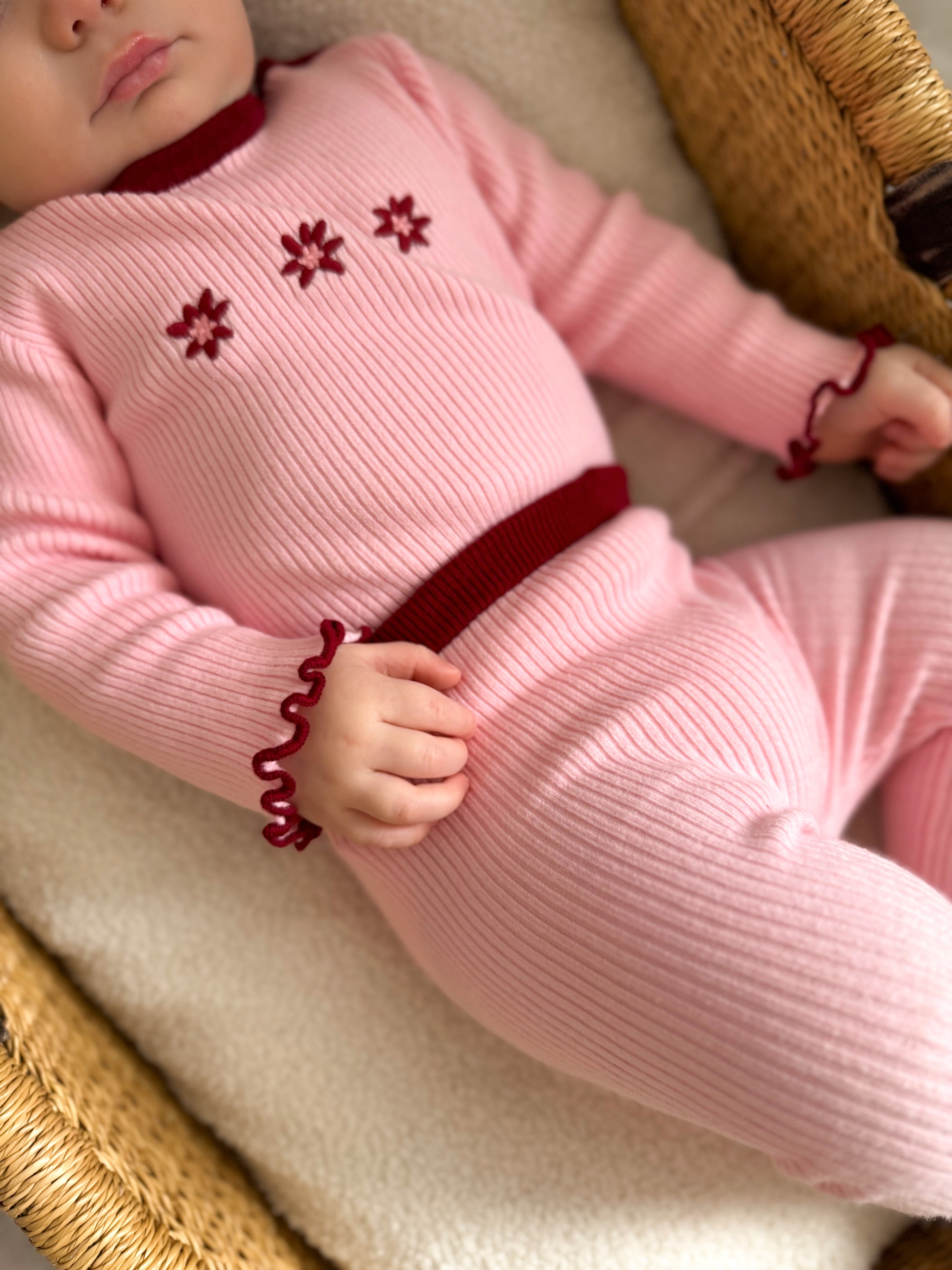 A baby dressed in ZIGGY LOU’s Leggings Lola—pink ribbed knit leggings with red trims and flower details—lies in a woven basket on a soft, cream-colored blanket.