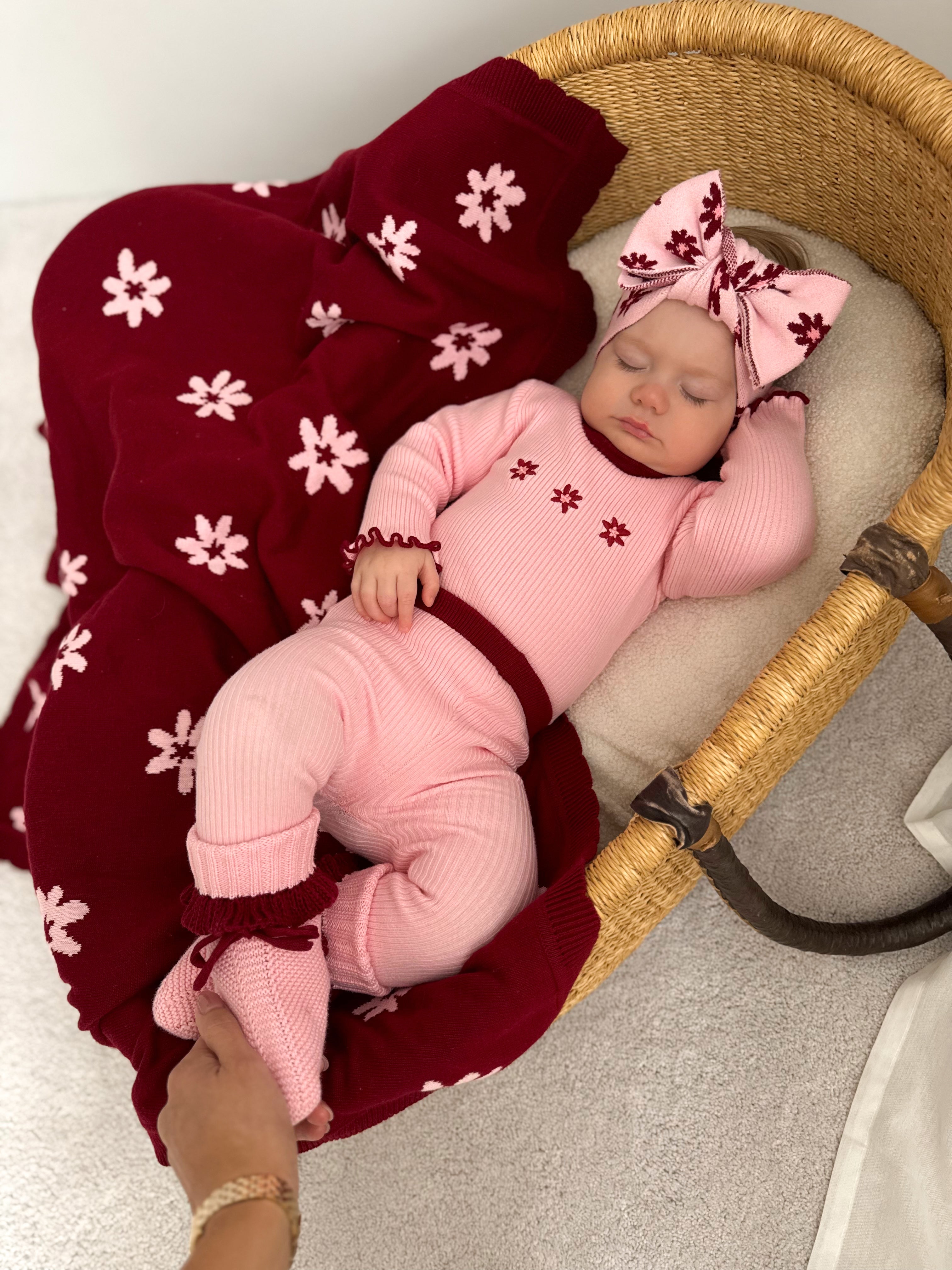 A baby in ZIGGY LOU - Leggings Lola with a matching bow headband sleeps in a wicker bassinet lined with a maroon, white-floral blanket as an adult hand gently holds the baby’s foot.