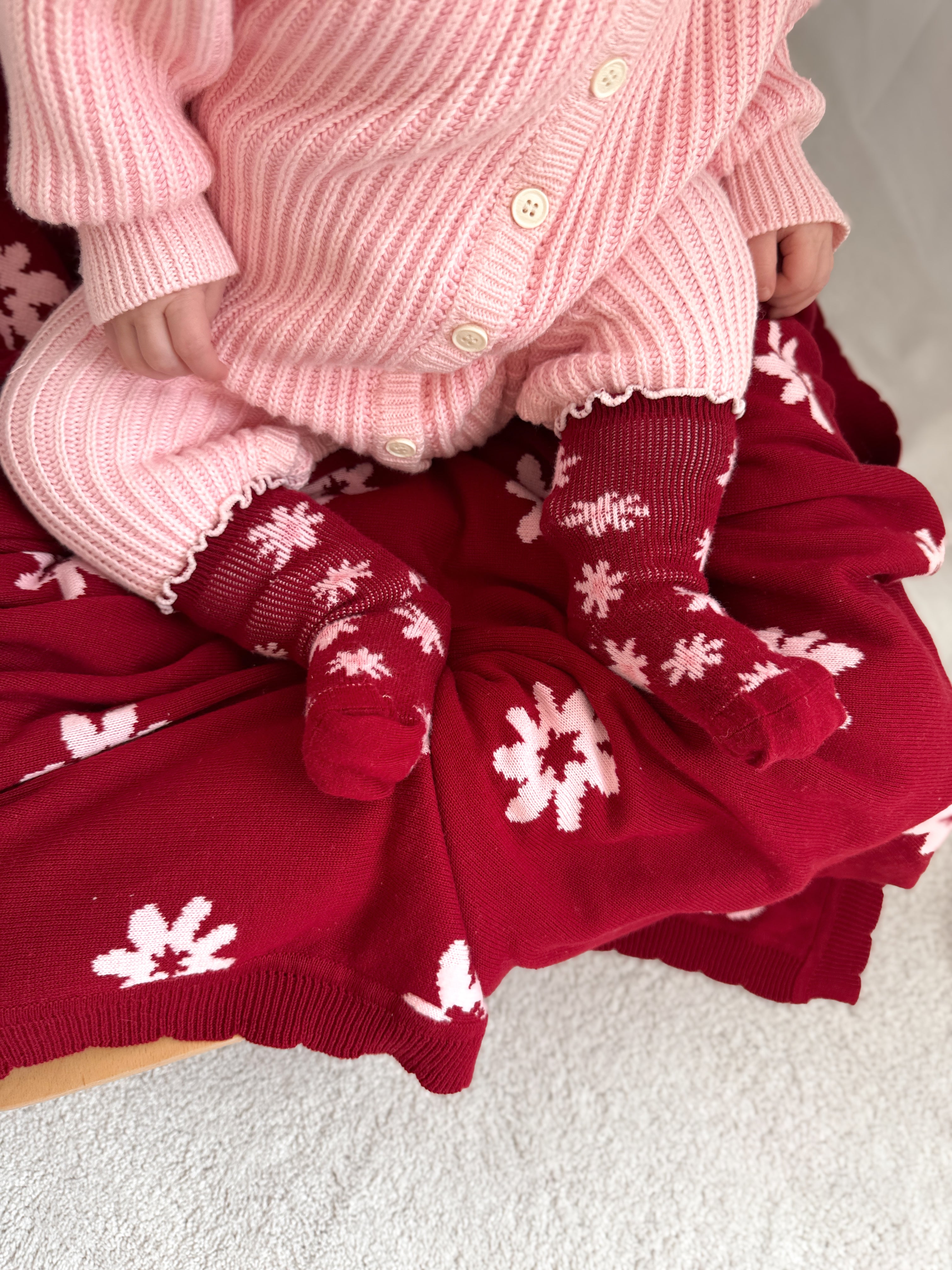 A baby in a pink ribbed outfit sits on a red Lola floral blanket, wearing ZIGGY LOU Ziggy Lou - Socks Lola (Plum) with white flower patterns. The baby's hands and legs are visible.