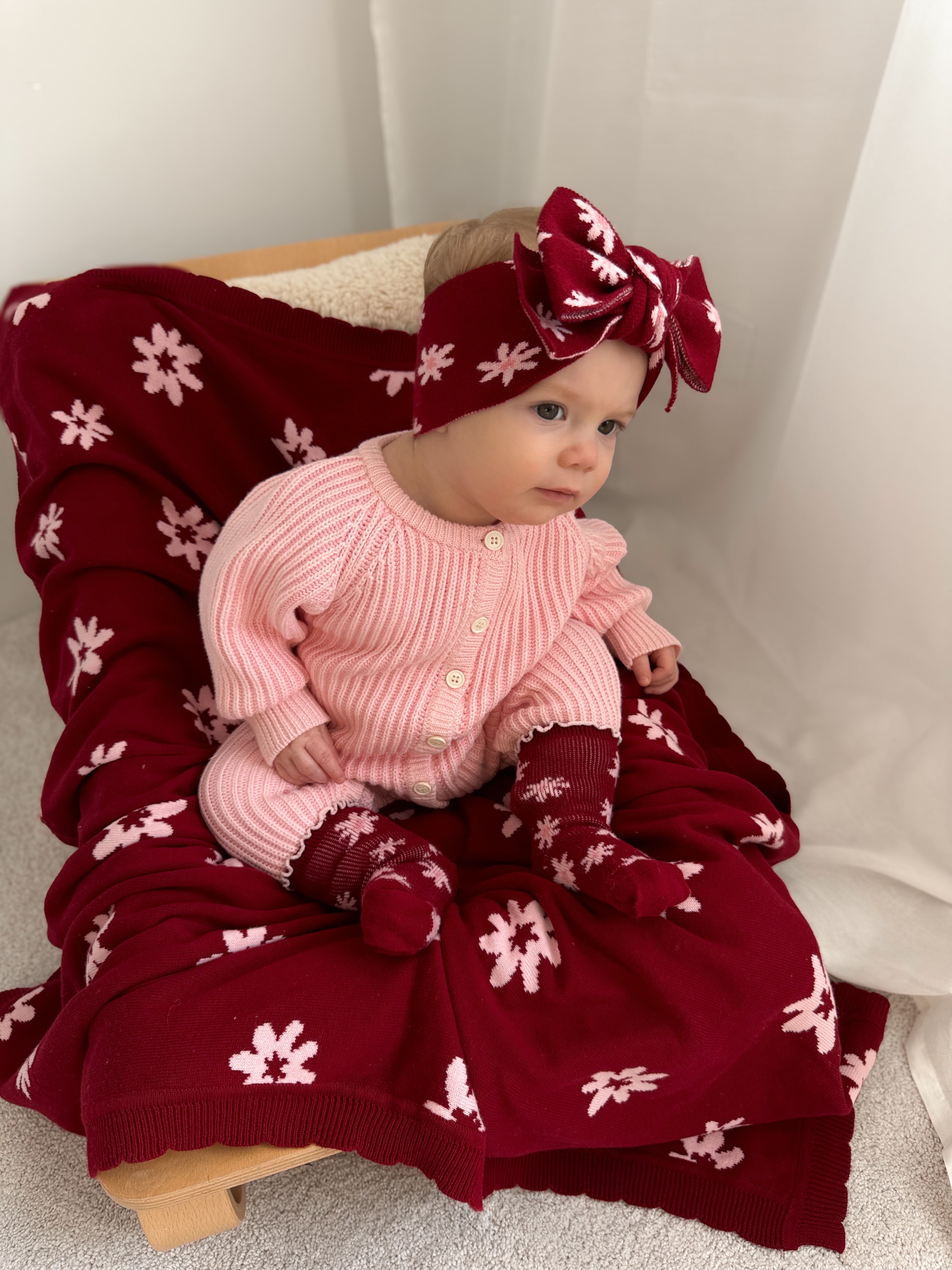 A baby in pink Ziggy Lou frill hem socks (Lola, Plum) by ZIGGY LOU, with a headband and large bow, sits on a maroon Lola floral blanket draped over a wooden chair.