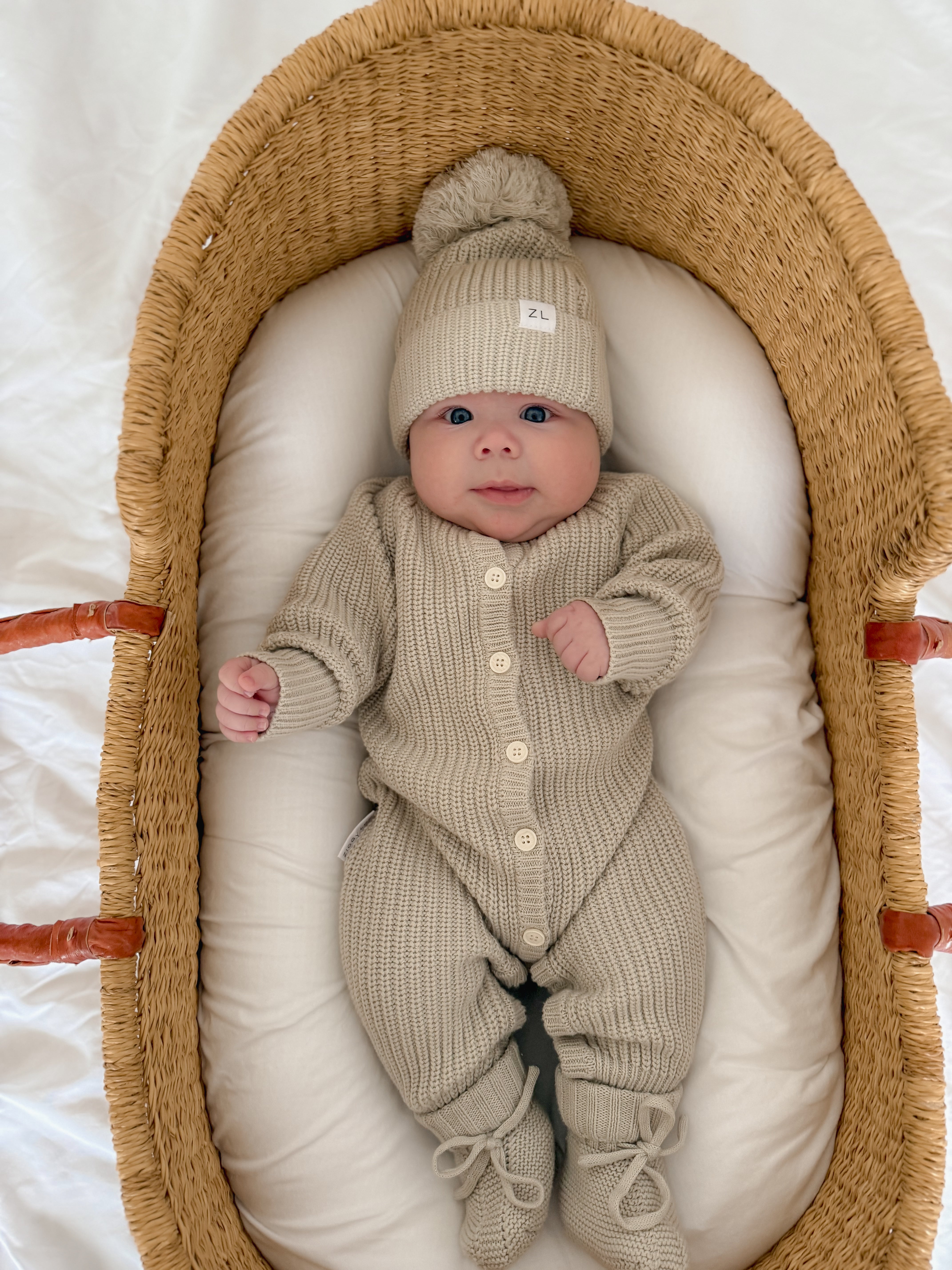 A baby relaxes in a woven basket on a white cushion, dressed in the ZIGGY LOU Ziggy Lou - Classic Knit Romper Moss with matching booties and a pom-pom beanie, gazing up wide-eyed.