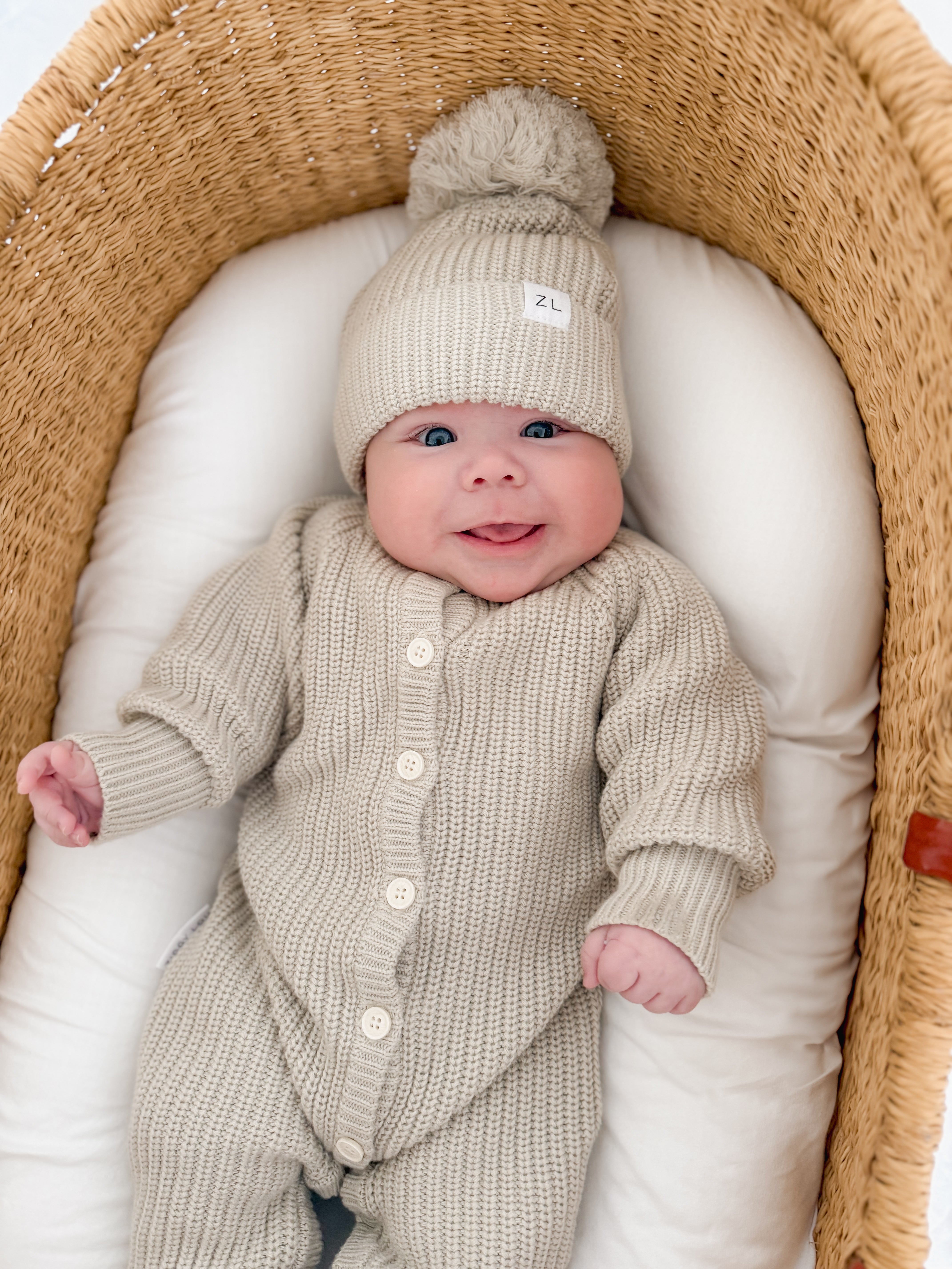 A smiling baby wears the Ziggy Lou - Classic Knit Romper Moss by ZIGGY LOU, lying in a wicker bassinet with a white cushion.