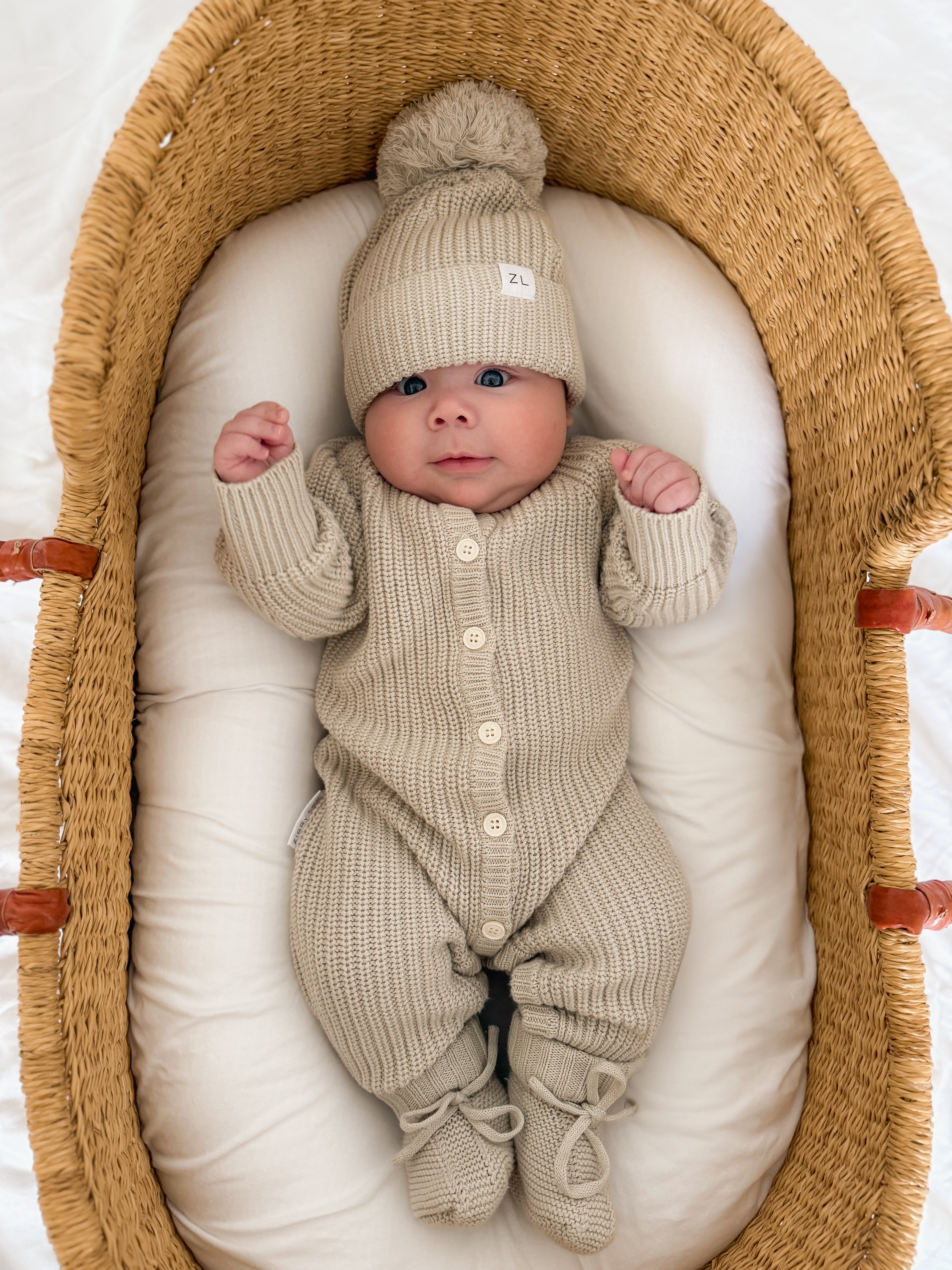 A baby in a ZIGGY LOU Ziggy Lou - Classic Knit Romper Moss lies on a white cushion inside a woven basket, looking up with hands raised.