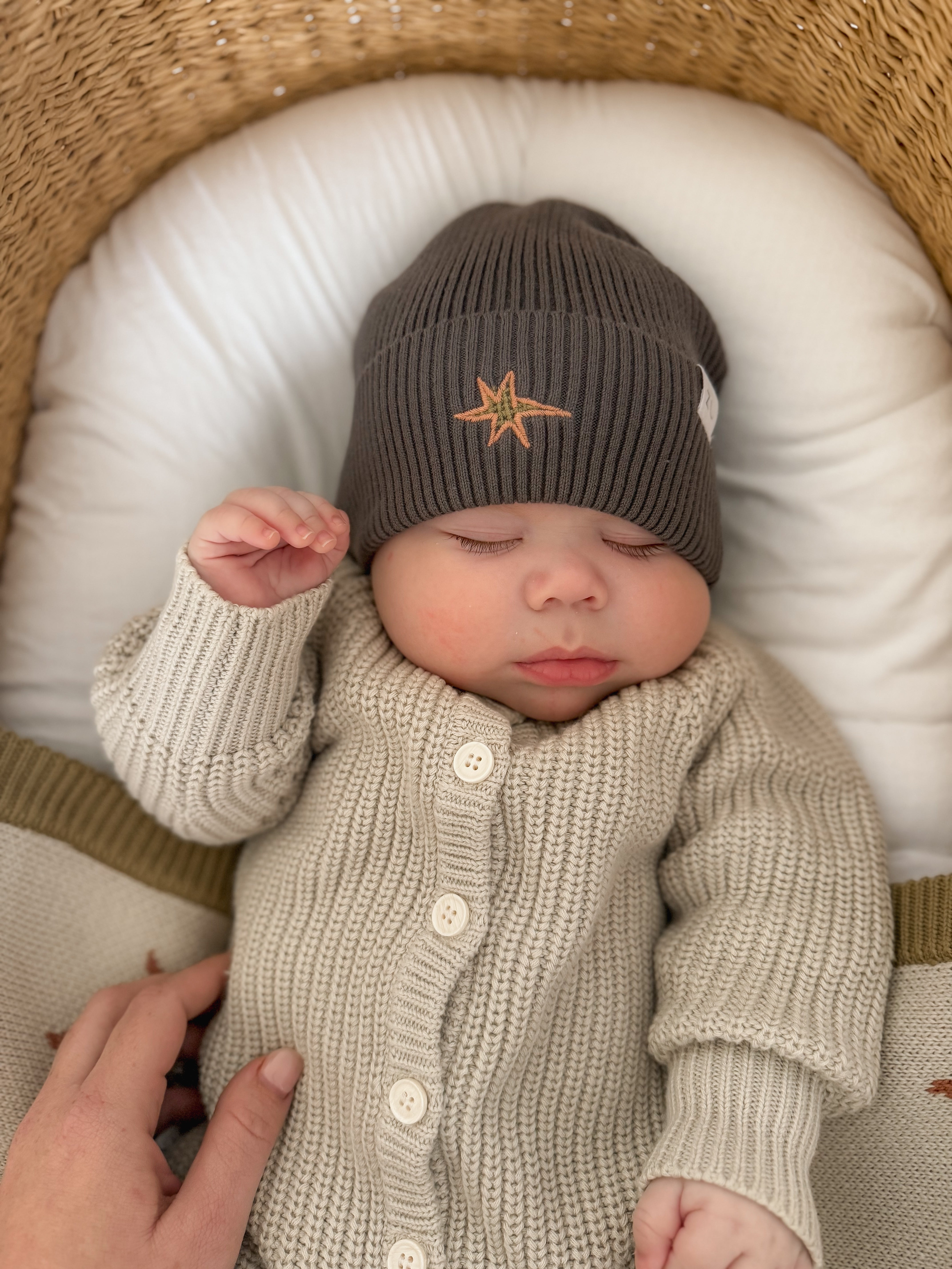 A baby sleeps in a woven bassinet, wearing the ZIGGY LOU Ziggy Lou - Classic Knit Romper Moss and a dark gray beanie with a star emblem. An adult hand gently touches the baby's side.