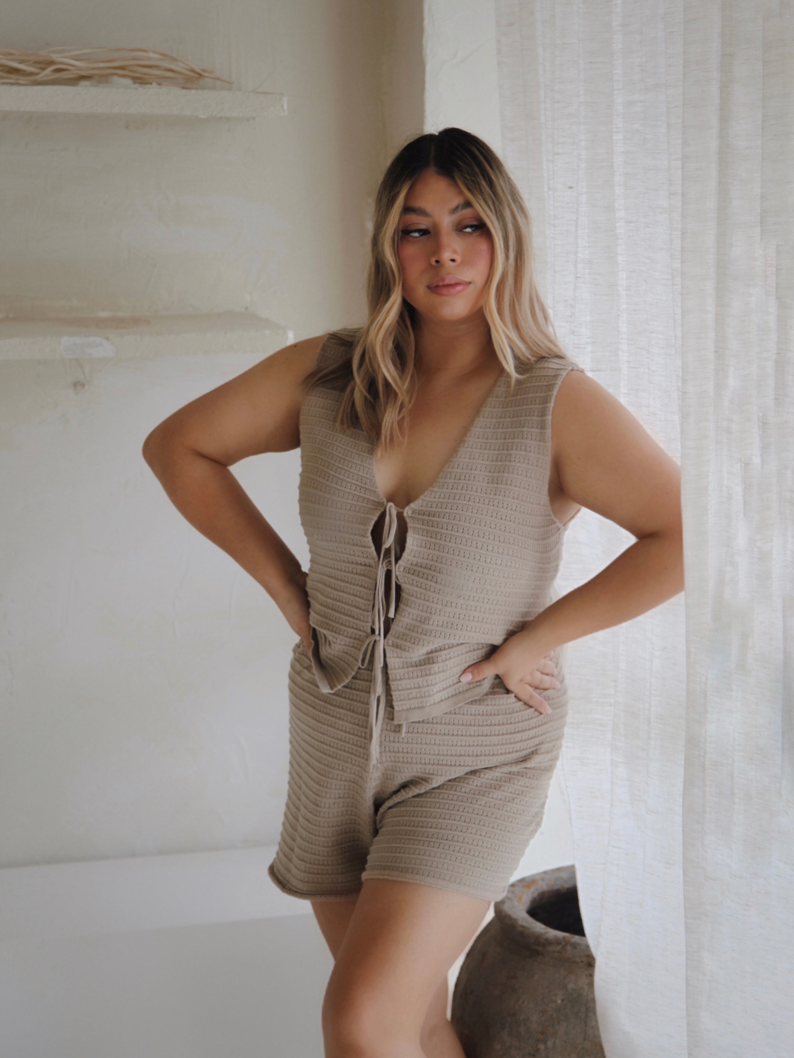A woman with long blonde hair stands by a window, wearing the Women's Knit Singlet Milo and matching shorts from ZIGGY LOU. This beige knitted eyelet singlet is crafted from organic cotton and features adjustable ties for a customized fit. The room has neutral-toned walls and decor, with soft outdoor light filtering through the curtain.