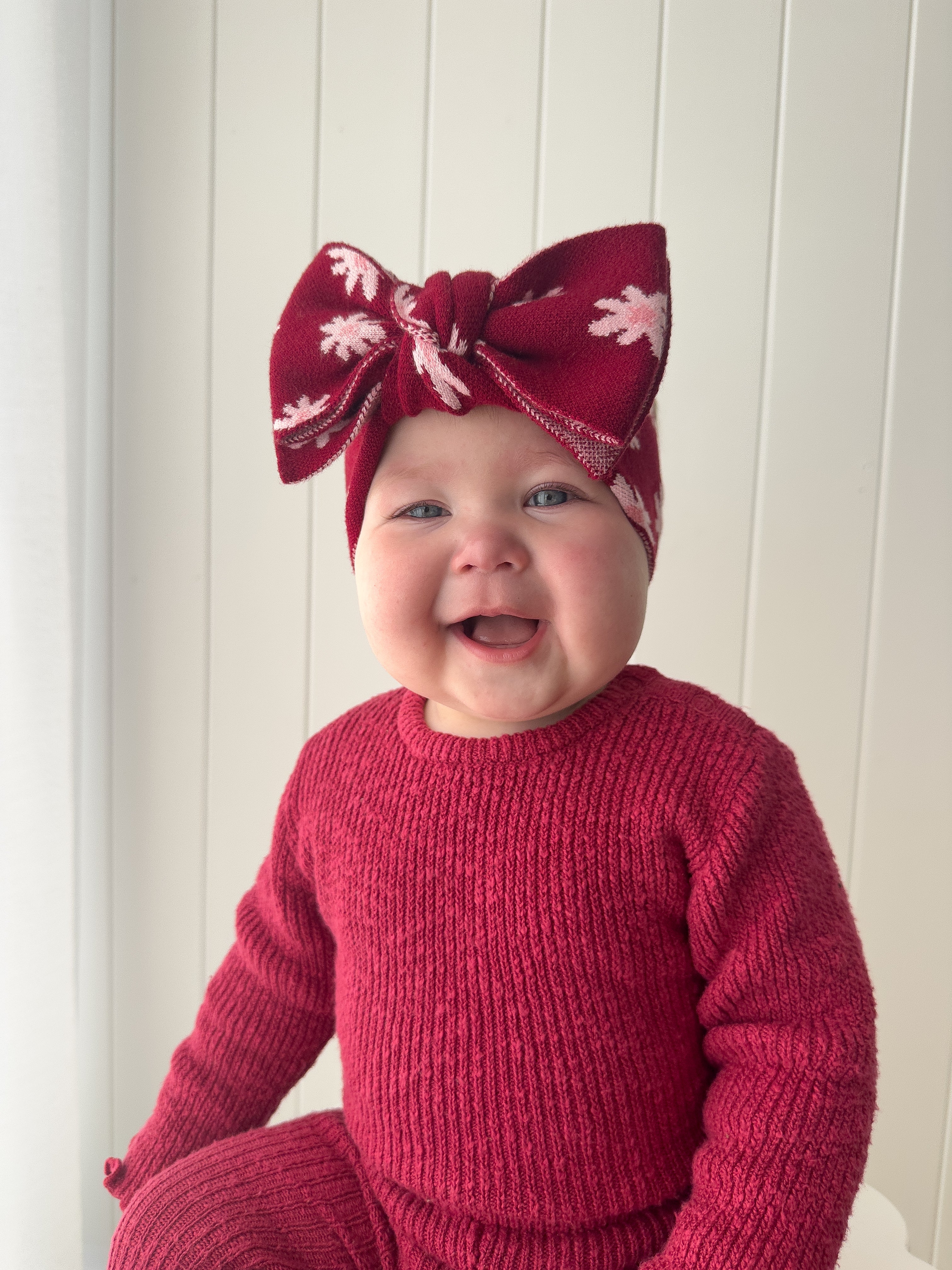 A smiling baby in a matching deep red knit outfit wears the ZIGGY LOU - Headband Lola (Plum), sitting in front of a white paneled wall.